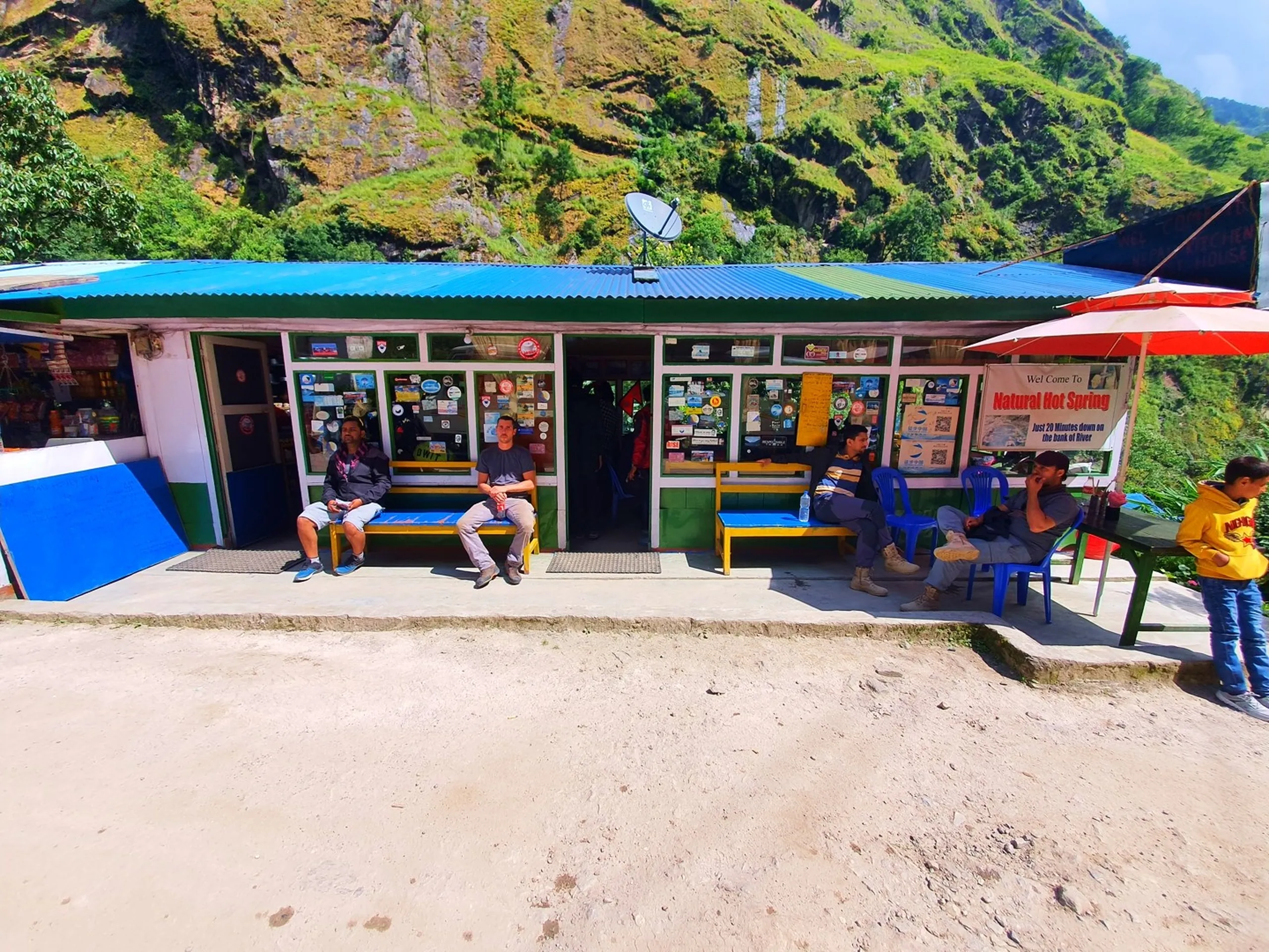 A small roadside teahouse and checkpoint in the Annapurna region of Nepal with a blue corrugated metal roof, green-trimmed windows plastered with stickers and notices, and a prominent banner reading 'Welcome to Natural Hot Spring - Just 20 Minutes Down on the Bank of River' - several trekkers and locals sitting on yellow benches and blue plastic chairs outside resting - a satellite dish on the roof, steep moss-covered cliffs rising directly behind the building - a typical Nepal trekking trail stop and natural hot spring signpost on the Annapurna Circuit, valuable content for Nepal hot spring trekking itinerary blogs