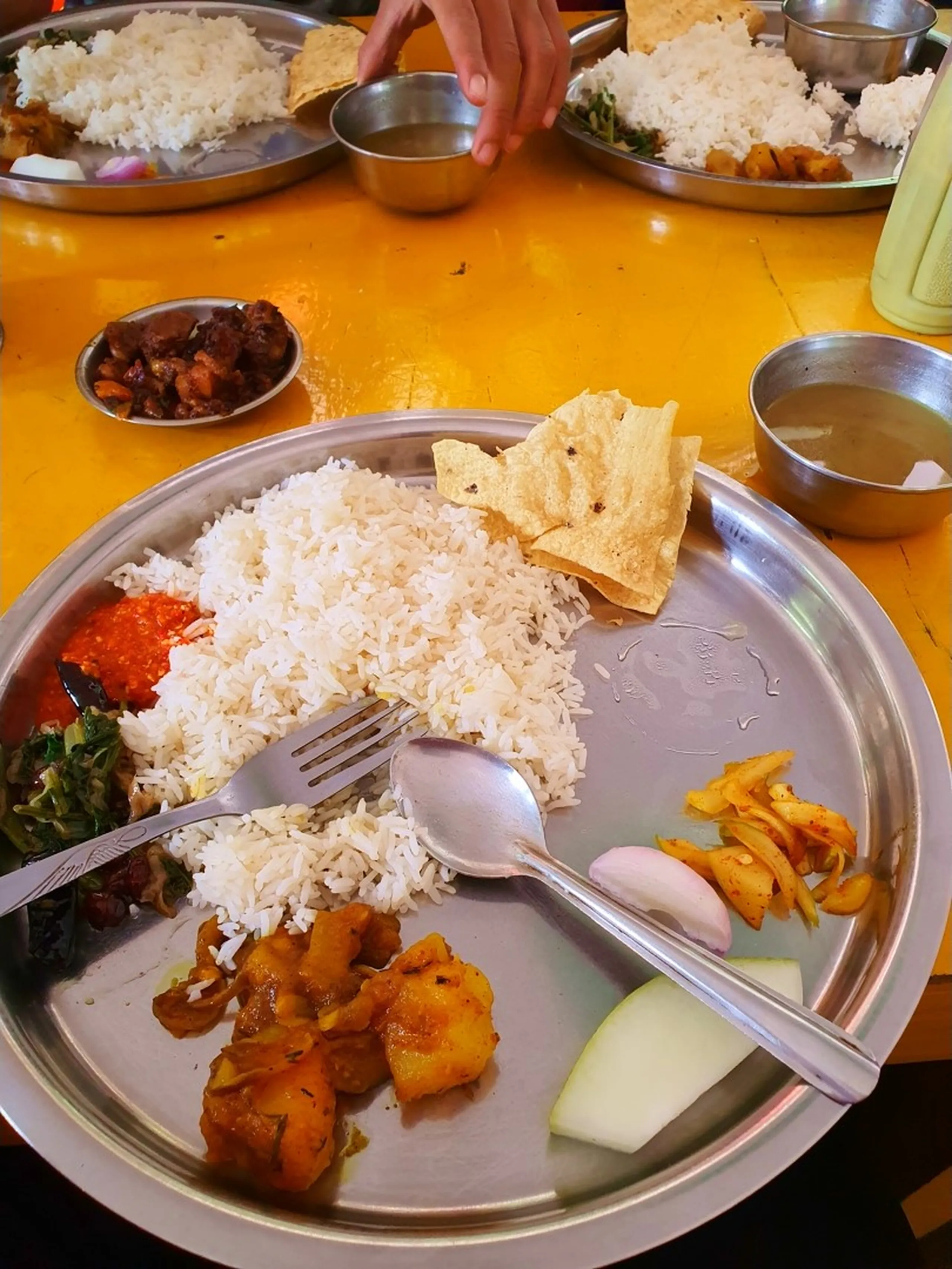 A close-up overhead shot of a traditional Nepali dal bhat meal served on a large round steel thali plate on a bright yellow table - steamed white rice piled in the center with a crispy papadum, surrounded by spiced potato tarkari curry, red chilli achar, sautéed leafy greens, sliced onion and cucumber, and a steel bowl of thin lentil dal soup - fork and spoon resting on the rice - additional thali plates and a green thermos visible in the background - an authentic Nepali trekking food photograph from a local teahouse on the Annapurna Circuit route to Manang, ideal for Nepal food blog and trekking meal content