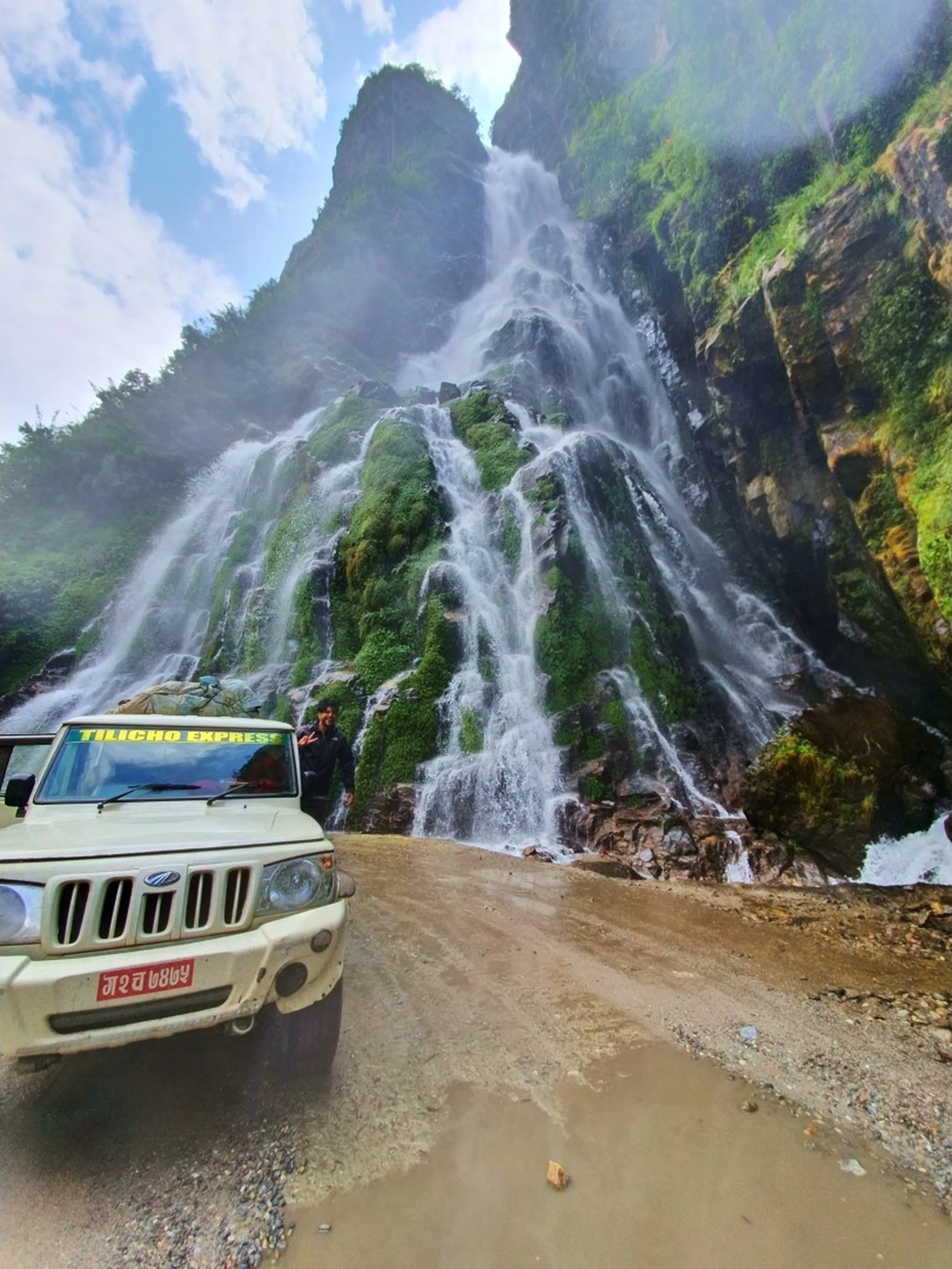 A breathtaking low-angle photograph of a massive multi-tiered waterfall cascading down a towering moss-covered rock face on the Annapurna Circuit road in Nepal - a white Mahindra Bolero jeep labeled 'Tilicho Express' parked in the foreground on a muddy mountain track, dwarfed by the enormous scale of the waterfall - a lone figure standing near the cliff face - the waterfall spreads across multiple green moss-draped ridges before crashing to the road level - dramatic clouds and jagged rocky peaks visible above - one of the most spectacular Nepal roadside waterfalls on the route to Manang, a must-see Annapurna Circuit attraction