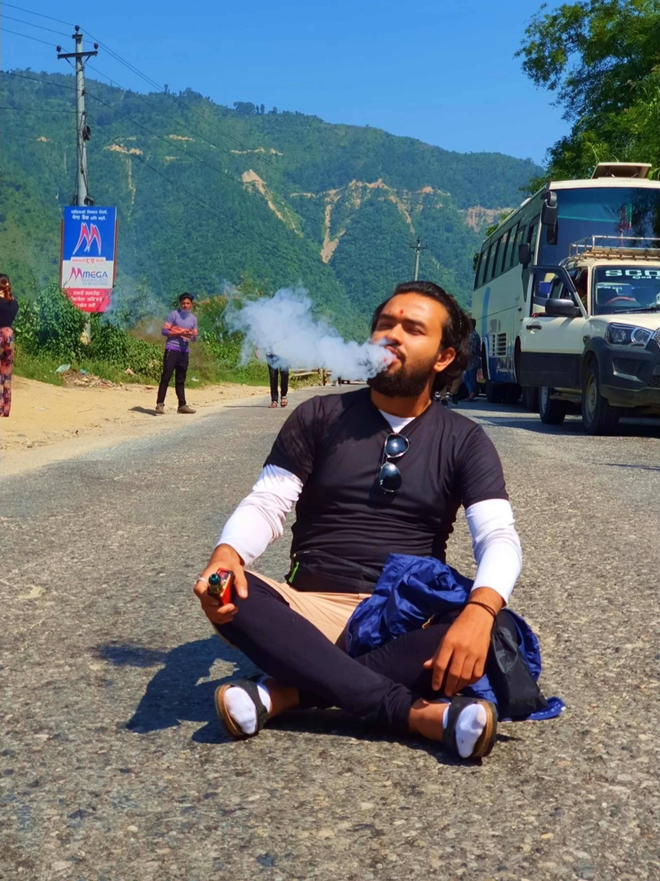 A young male trekker sits cross-legged in a meditation-like pose in the middle of a mountain highway in Nepal, vaping with a large cloud of smoke drifting skyward, wearing a black long-sleeve layered shirt with white undershirt, sunglasses hanging from his collar, a blue jacket draped on his lap, parked buses and a white SUV behind him, lush green terraced hills and a Mega Bank billboard visible in the background - a candid road trip moment during a Nepal trekking expedition to Manang
