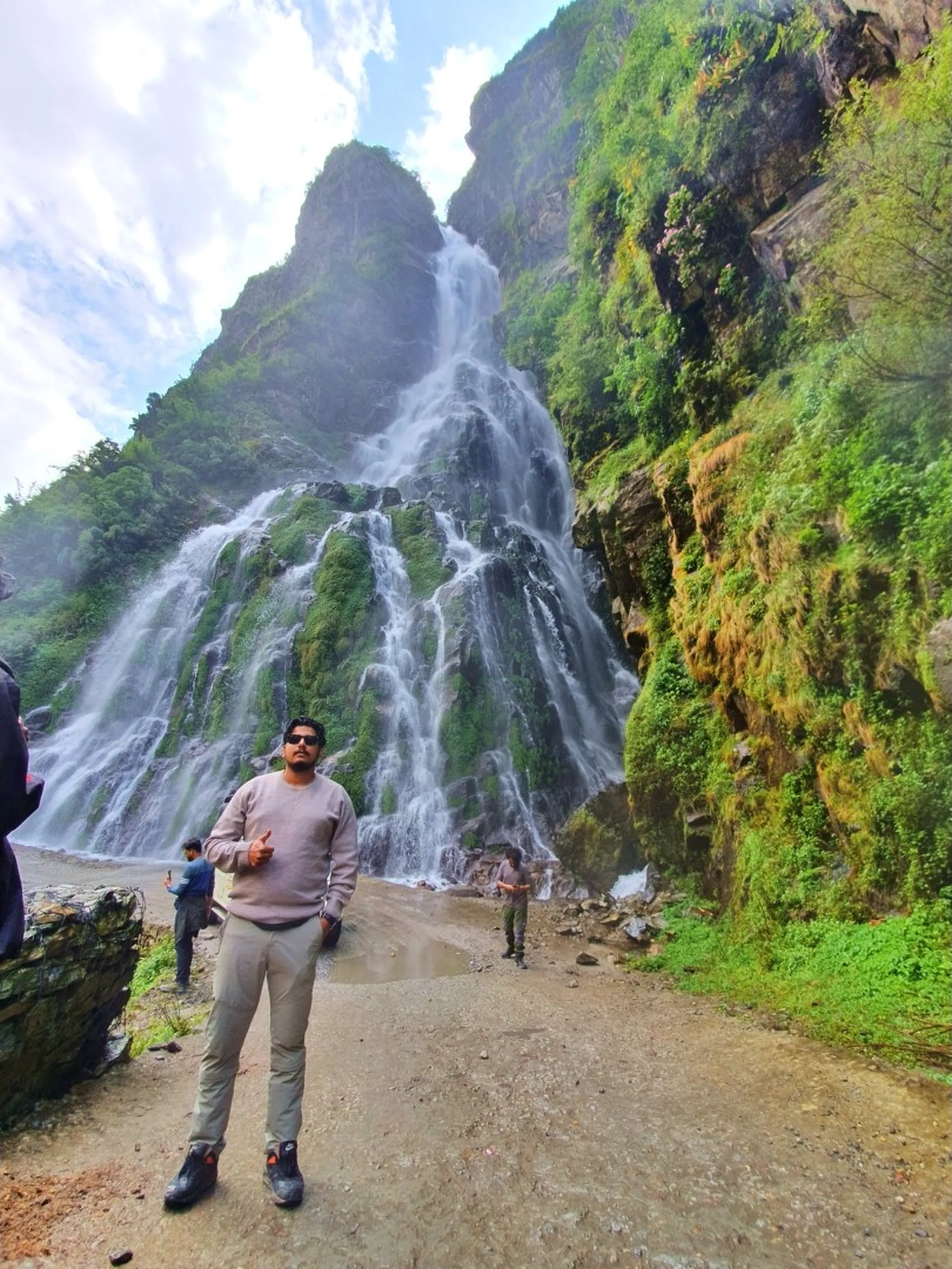 A young male Nepali trekker wearing a dusty rose-pink sweatshirt, beige trekking trousers, black hiking boots, and dark sunglasses stands giving a shaka hand gesture in front of an enormous multi-stream waterfall on the Annapurna Circuit road to Manang - the waterfall fans out across a massive green moss-covered cliff face behind him, generating light mist - two other trekkers visible in the mid-ground near the base - a dramatic Nepal adventure portrait at one of the most beautiful roadside waterfalls in Nepal, on the Besisahar to Manang jeep route, Annapurna region