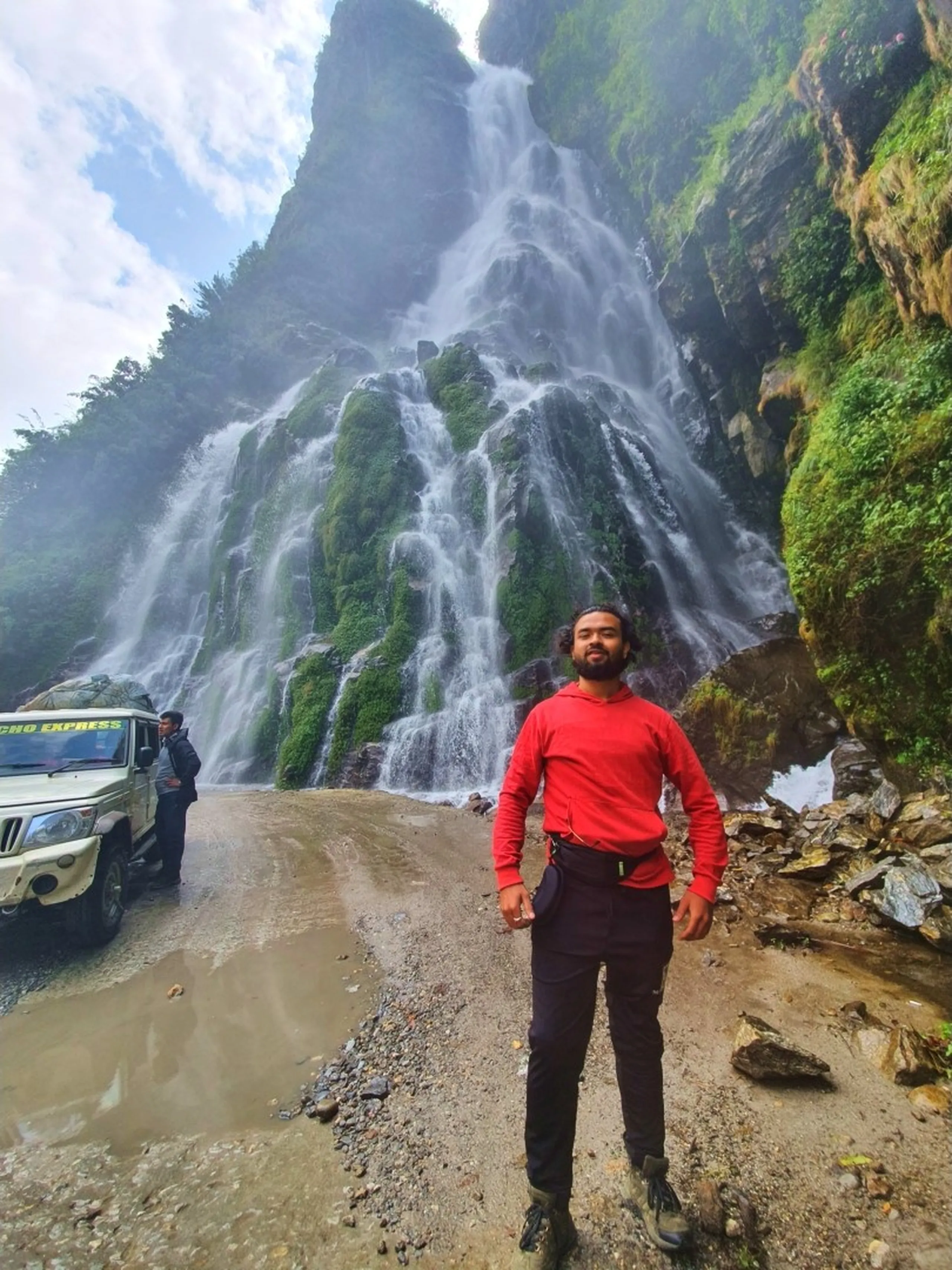A young male trekker in a bright red hoodie, black trekking pants, and hiking boots stands tall and composed in front of a colossal multi-cascade waterfall on a muddy mountain road in the Annapurna Circuit, Nepal - the waterfall tumbles down a towering green moss-layered cliff face in multiple white streams behind him - a white Tilicho Express jeep and another group member visible to the left - heavy mist surrounds the base - one of the largest and most dramatic Nepal roadside waterfalls on the Manang trek route, perfect for Nepal travel photography and Annapurna Circuit blog content