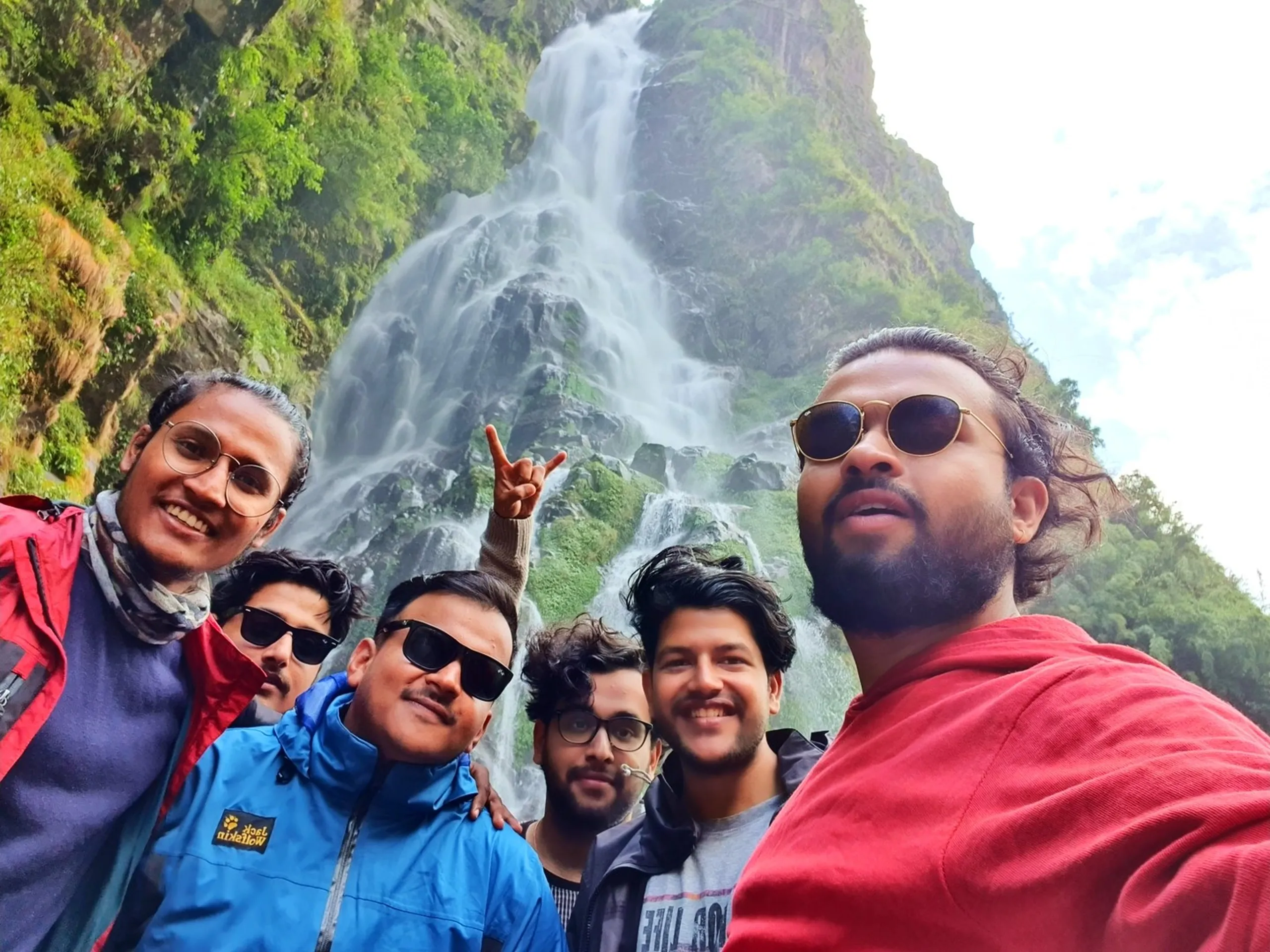 A group selfie of six young male Nepali trekkers taken directly in front of a spectacular multi-tiered waterfall on the Annapurna Circuit, Nepal - wearing jackets in red, blue, black, and grey tones, with various sunglasses and scarves - one flashing a rock hand sign, all grinning and laughing - the enormous white waterfall fans across a green moss-covered rocky cliff face filling the entire background - an exuberant Nepal trekking group photo capturing the joy and brotherhood of a Himalayan adventure, on the road to Manang, one of the best Nepal waterfalls along the Annapurna Circuit jeep route