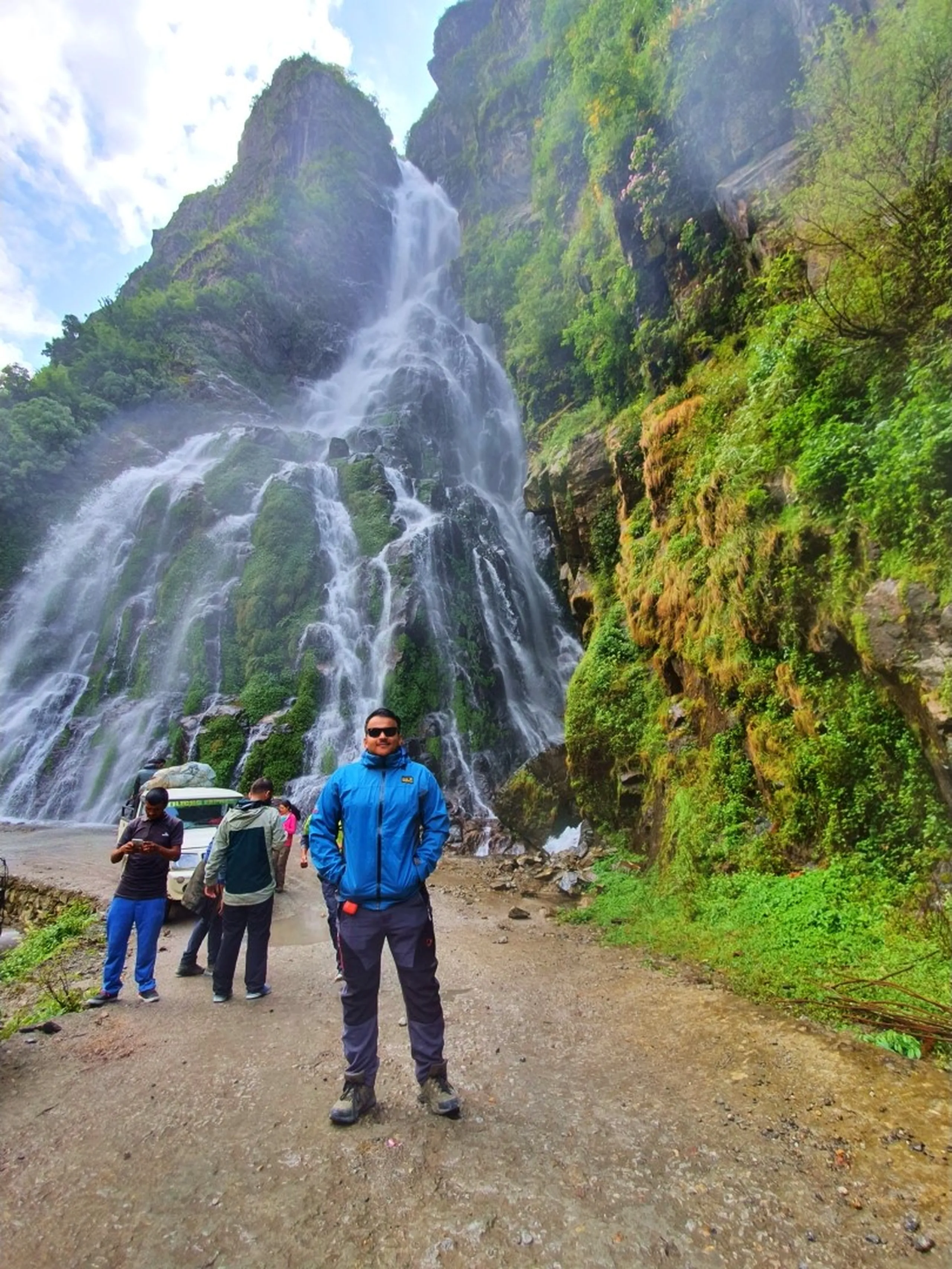 A confident young male trekker wearing a bright blue Jack Wolfskin trekking jacket, black trekking pants, and hiking boots stands hands-in-pockets in front of a thundering multi-cascade Nepal waterfall on the Annapurna Circuit - the massive waterfall spreads across a towering moss-drenched cliff behind him, white streams diverging over green rock - other group members and a parked jeep visible to the left - mist rising at the base - a stunning Nepal trekking portrait at one of the most famous waterfalls on the route from Besisahar to Manang, great for Annapurna Circuit travel blog imagery