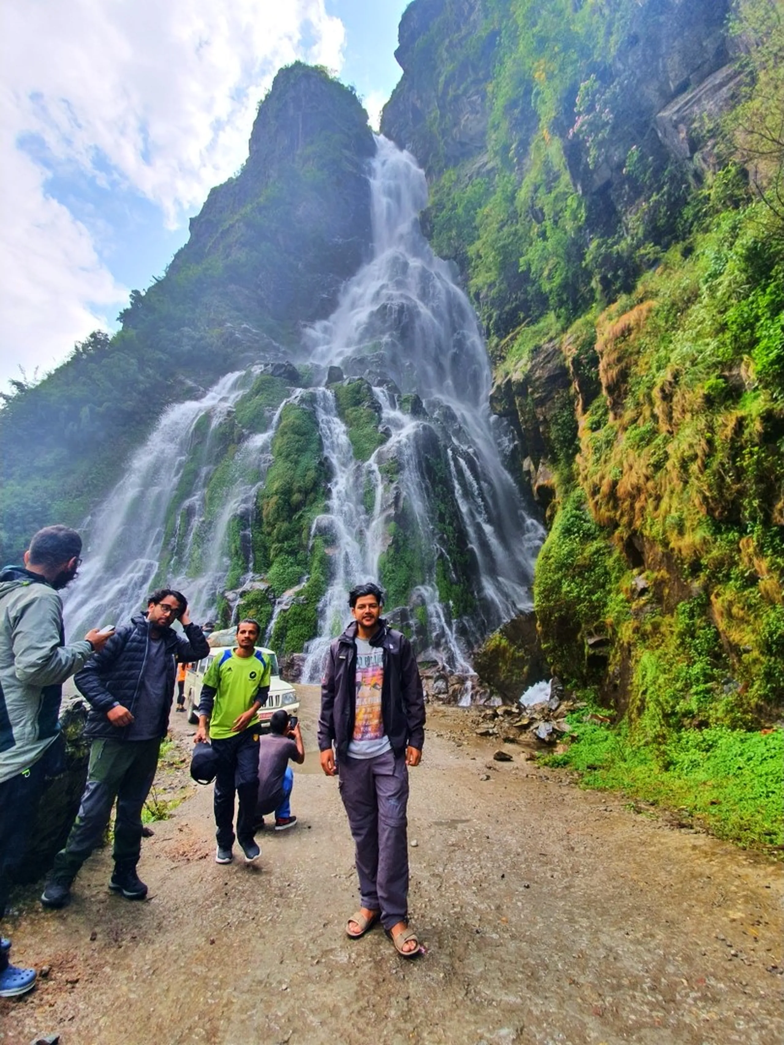 A slim young male trekker wearing a dark grey rain jacket, grey graphic tee, grey trousers, and sandals stands in the center foreground of a dramatic Nepal waterfall scene on the Annapurna Circuit - the massive multi-stream waterfall cascades down a towering green moss-covered cliff face behind him - fellow trekkers in the background including one in a neon green jacket photographing the waterfall - parked jeep visible at the road edge - misty atmosphere with jagged rocky peaks above - a candid Nepal trekking portrait at one of the most awe-inspiring roadside waterfalls on the Manang route, capturing the raw scale of Himalayan nature