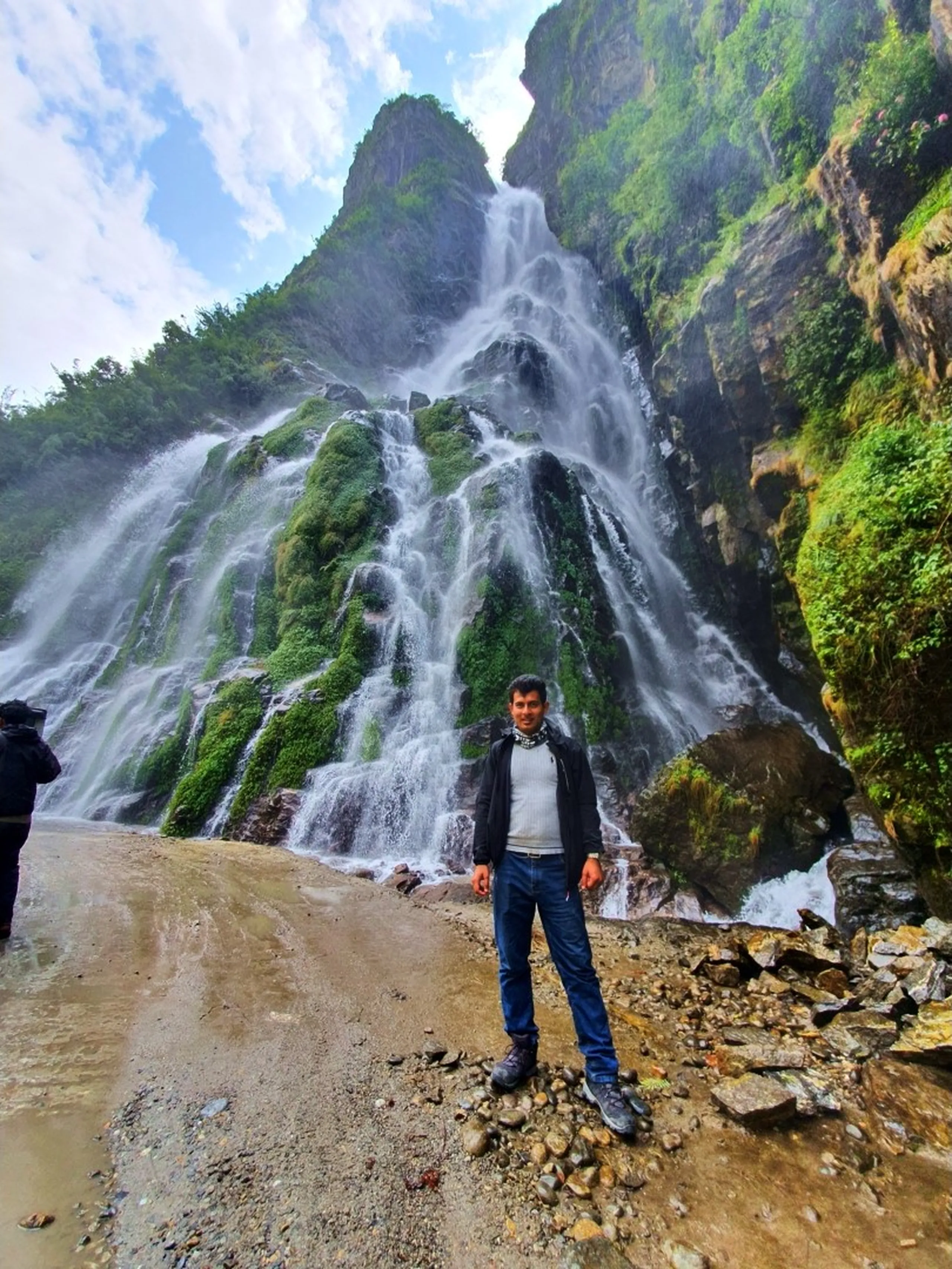 A young male trekker wearing a grey jacket layered over a white turtleneck, blue jeans, and hiking boots stands centered in front of a massive multi-cascade Nepal waterfall with arms relaxed - the waterfall explodes across a huge green moss-covered rock face filling the background wall-to-wall, mist rising at the base - rocky and muddy ground in the foreground - another trekker partially visible on the left edge - a powerful and immersive Nepal waterfall photography portrait on the Annapurna Circuit jeep road to Manang, showcasing the incredible scale of Himalayan waterfalls Nepal is known for