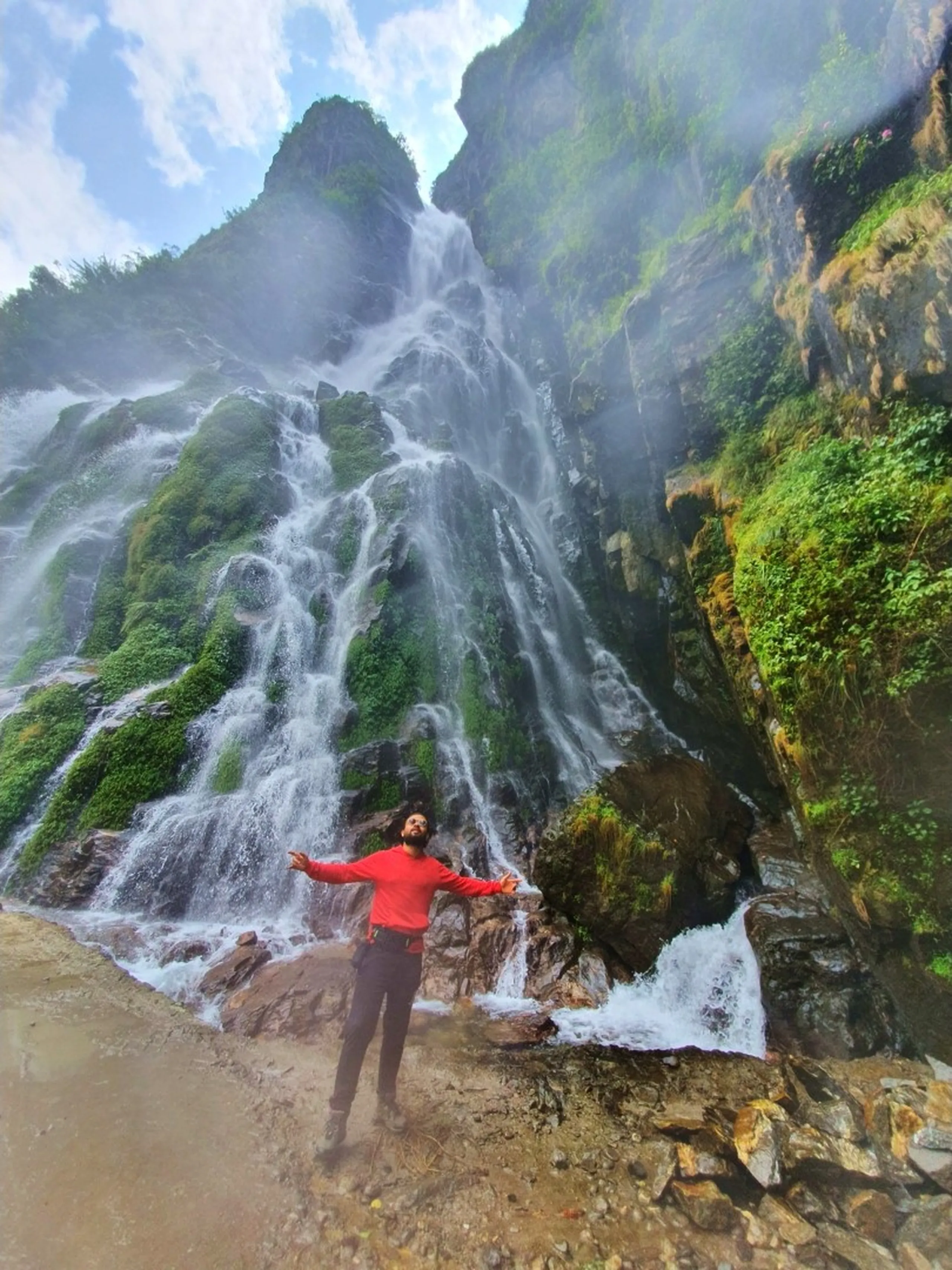 A young male trekker in a bright red hoodie stands at the base of a colossal waterfall with arms stretched wide open in a gesture of pure freedom and awe - the waterfall surges down in multiple white torrents over a towering green moss-covered rocky cliff that dominates the entire frame - mist swirls dramatically around him - standing on muddy rocky ground at the road level - a wide-angle low perspective amplifies the scale and power of the water - an iconic Nepal adventure travel photograph at one of the most spectacular waterfalls on the Annapurna Circuit, Manang trekking route, Nepal, capturing the spirit of Himalayan exploration