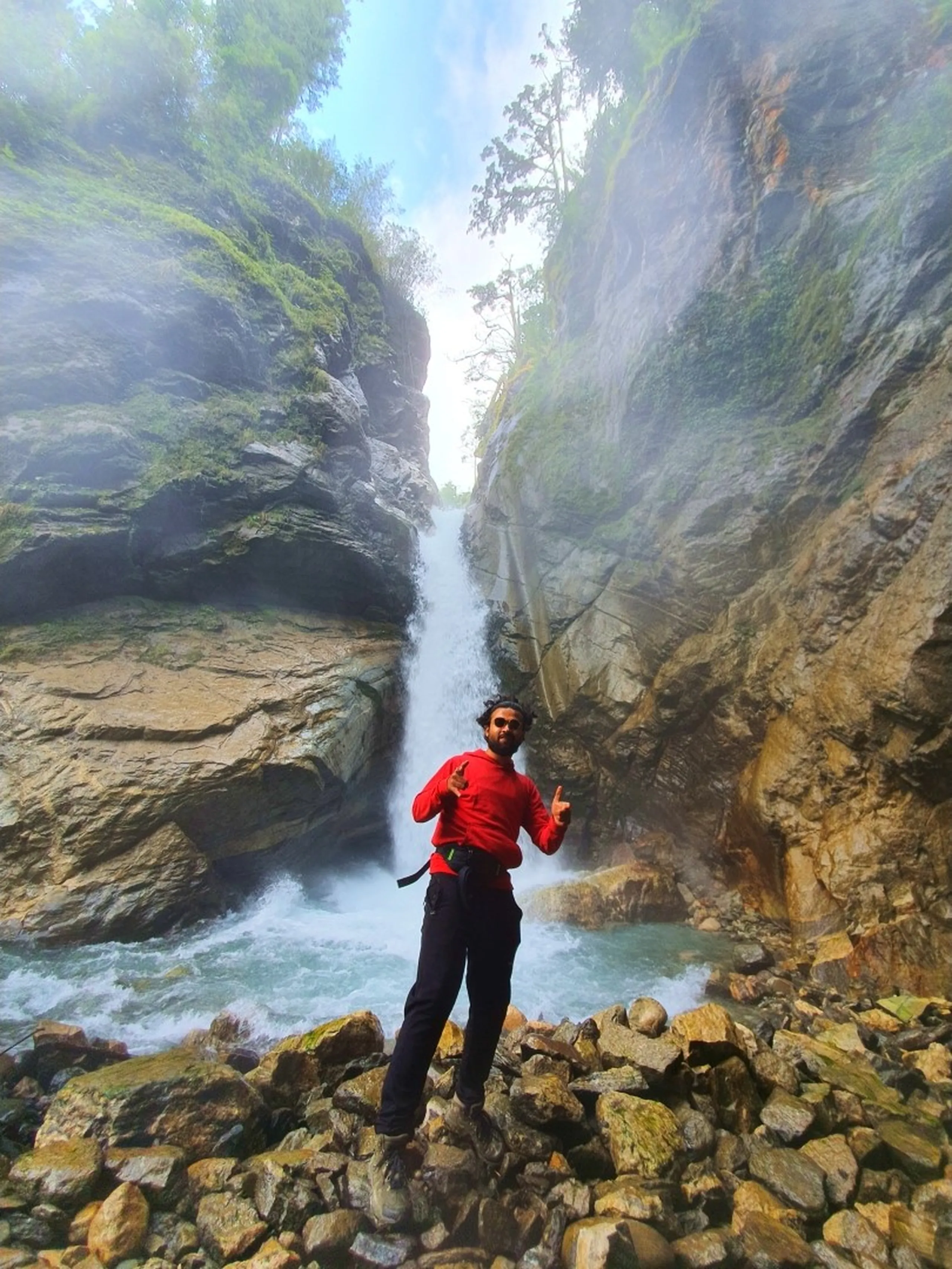 A young male trekker in a bright red hoodie and black trekking pants stands triumphantly on a pile of mossy boulders at the base of a deep narrow gorge waterfall in Nepal - the waterfall plunges from a dramatic rocky slot canyon between two towering cliff walls, turquoise water churning at the base - misty spray fills the air - the trekker flashes double finger-gun poses with a beaming smile - dense green vegetation clings to the cliff tops above - an exhilarating Nepal gorge waterfall photograph on the Annapurna Circuit trail between Besisahar and Manang, capturing the hidden canyon waterfall beauty of Nepal trekking