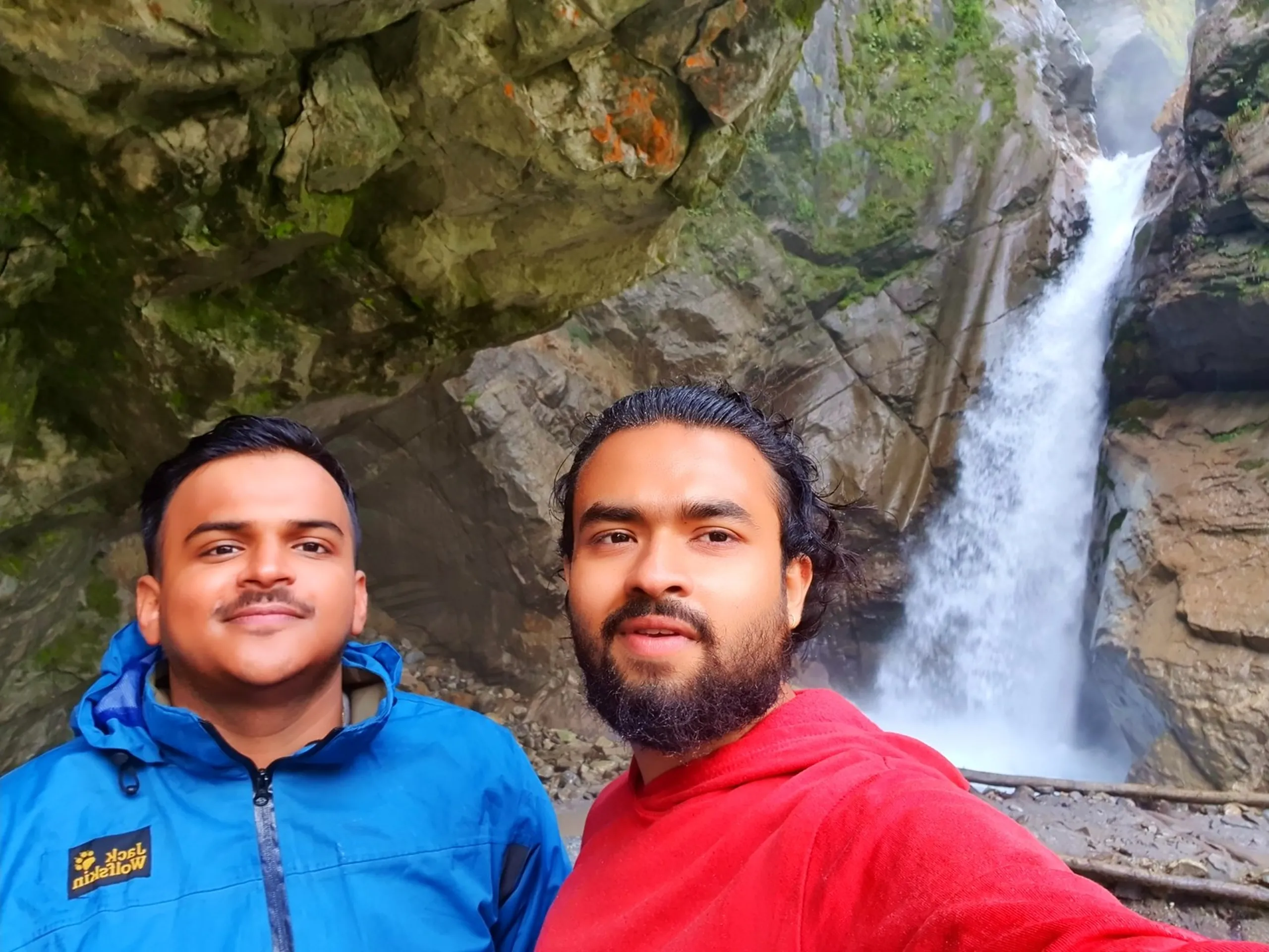 A close-up selfie of two young male Nepali trekkers at the base of a dramatic gorge waterfall in Nepal - one wearing a blue Jack Wolfskin jacket with a neatly styled haircut and the other in a red hoodie with shoulder-length hair tied back and a full beard - both smiling warmly at the camera - the waterfall plunges through a narrow rocky canyon slot behind them, white water thundering between tall stone walls - a candid Nepal trekking friendship portrait inside a hidden gorge waterfall on the Annapurna Circuit route to Manang, showcasing the bond formed on long Himalayan treks