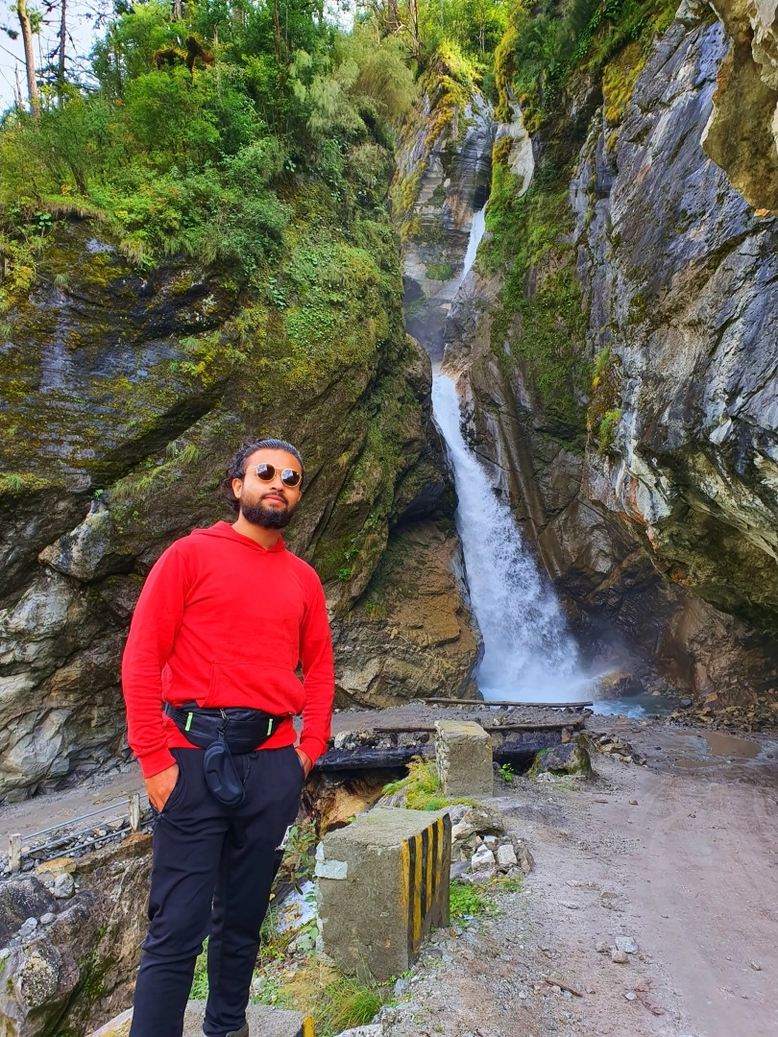 A young male trekker in a bright red hoodie, black trekking pants, fanny pack, and round mirrored sunglasses poses confidently beside a dramatic single-plunge waterfall that drops through a narrow rocky canyon gorge in Nepal - the waterfall falls steeply between two massive grey rock walls draped in green moss and vegetation - a small concrete road barrier with black-and-yellow chevron stripes sits in the foreground - the road and a small metal bridge at the waterfall base visible in the distance - a striking Nepal canyon waterfall portrait on the Annapurna Circuit road between Lunjung and Manang, showcasing the rugged dramatic landscape of trekking in Nepal