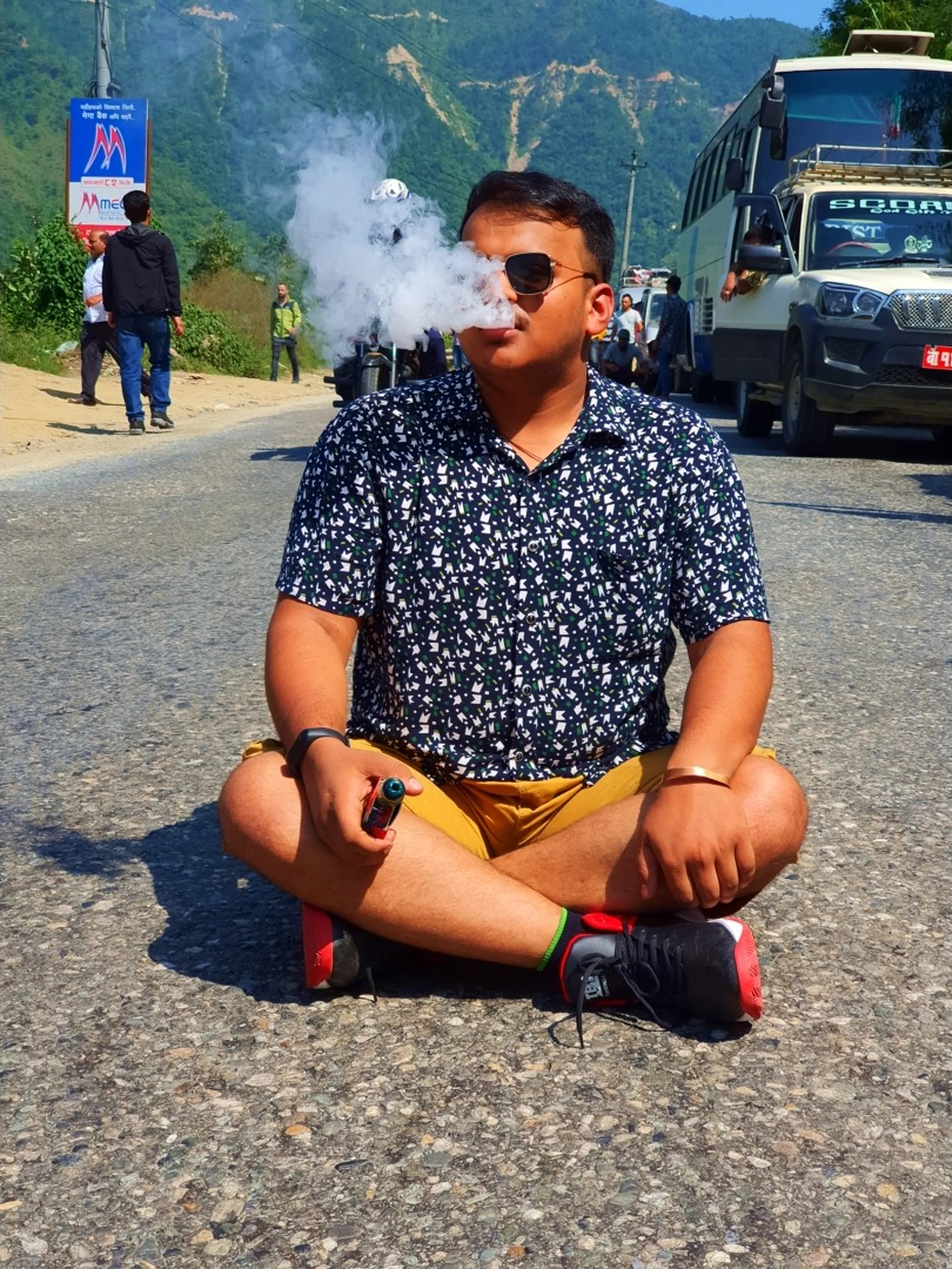 A young male trekker sits cross-legged on a mountain road in Nepal exhaling a massive vape cloud, wearing a bold patterned navy short-sleeve shirt, yellow cargo shorts, black and red sneakers, and aviator sunglasses, holding a lighter in one hand - traffic jam backdrop with parked buses, SUVs, and green Himalayan hills stretching across the horizon under a clear blue sky, capturing the raw and spontaneous spirit of Nepal road trip adventures