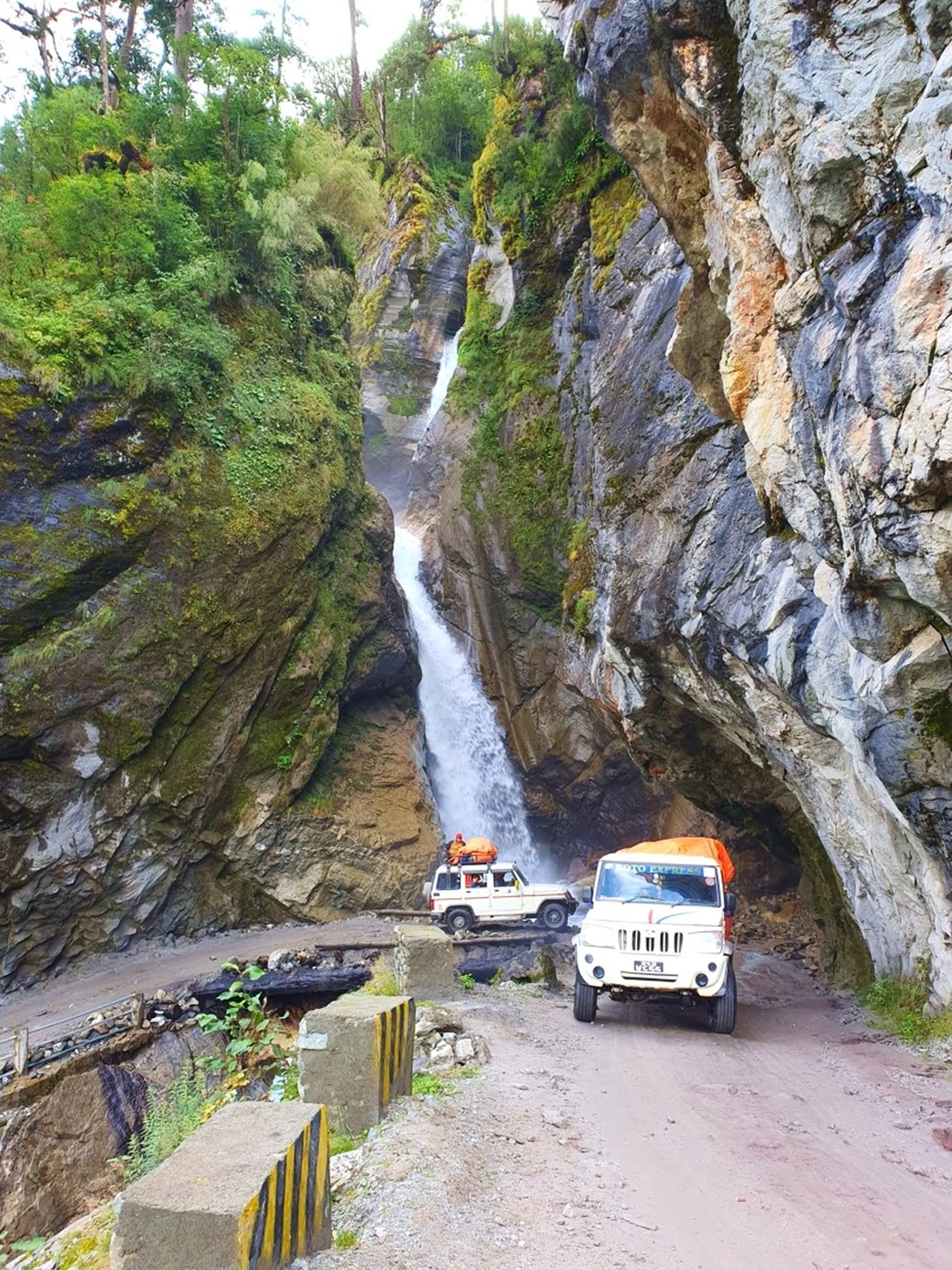 A wide-angle aerial-perspective photograph of two white Mahindra Bolero 4WD jeeps - one labeled 'Tilicho Express' - navigating a narrow dirt mountain road inside a dramatic rocky gorge canyon in Nepal - a powerful single-drop waterfall plunges between two towering rock walls directly above and behind the vehicles, dwarfing them in scale - moss and green shrubs cling to the canyon walls, a small concrete bridge and road barrier visible - the scene conveys the extreme and thrilling terrain of the Annapurna Circuit jeep road between Lunjung and Manang, Nepal - a spectacular Nepal mountain road adventure photograph perfect for Annapurna Circuit travel guides and Nepal 4WD jeep trekking content