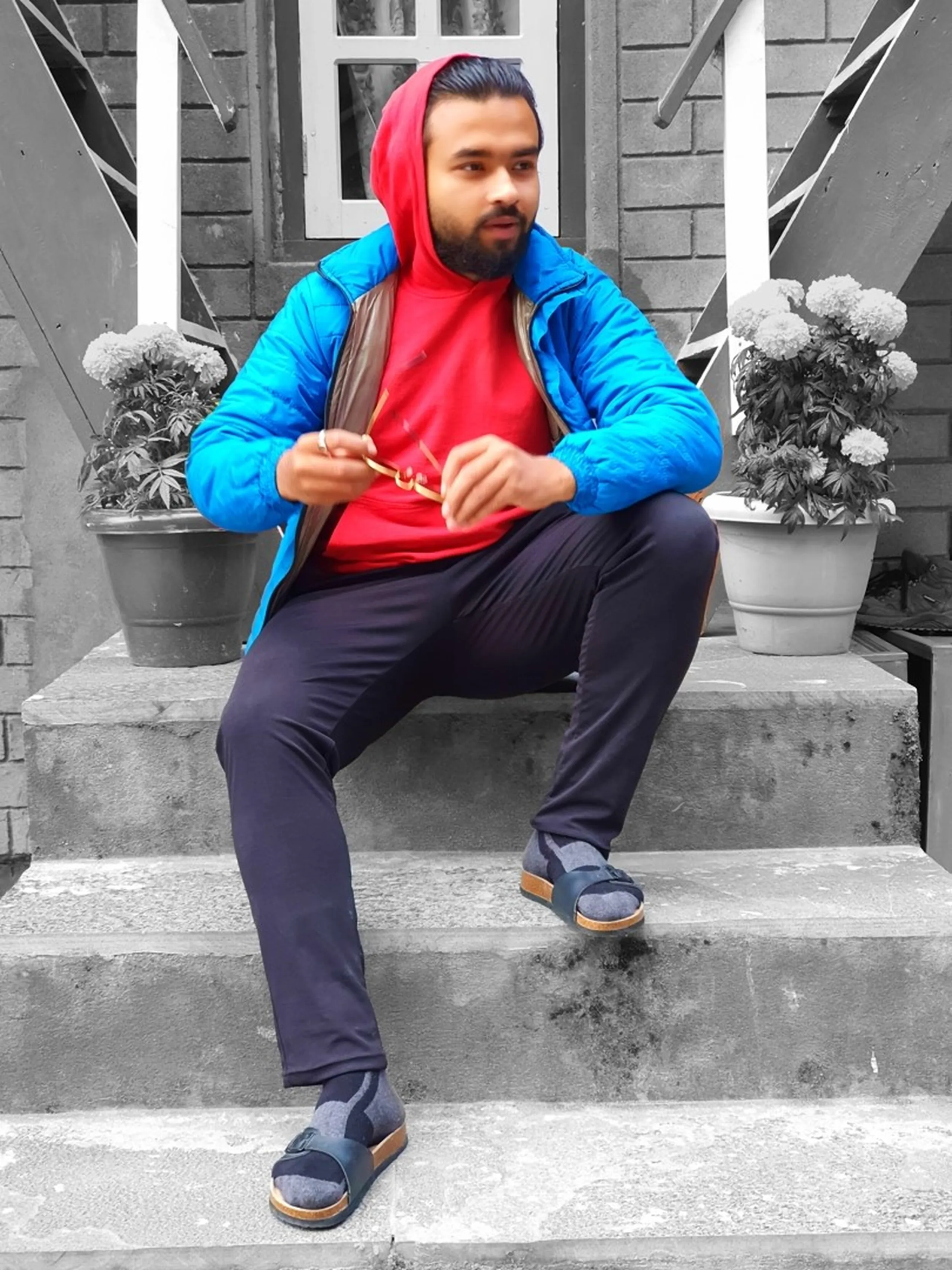 A creative selective-color portrait photograph of a young male Nepali trekker sitting casually on stone steps outside a guesthouse in Chame, Manang, Nepal - wearing a bright red hoodie under a vivid blue puffer jacket, dark trekking trousers, and grey open-toe sandals, holding gold-framed sunglasses in his hands - the entire background and surroundings are rendered in black and white, including potted flowering plants on either side and a stone-brick building facade with a white-framed window - only the subject's colorful clothing pops in full color - a stylish and artistic Nepal trekking lodge portrait, perfect for Annapurna Circuit lifestyle and travel blog content