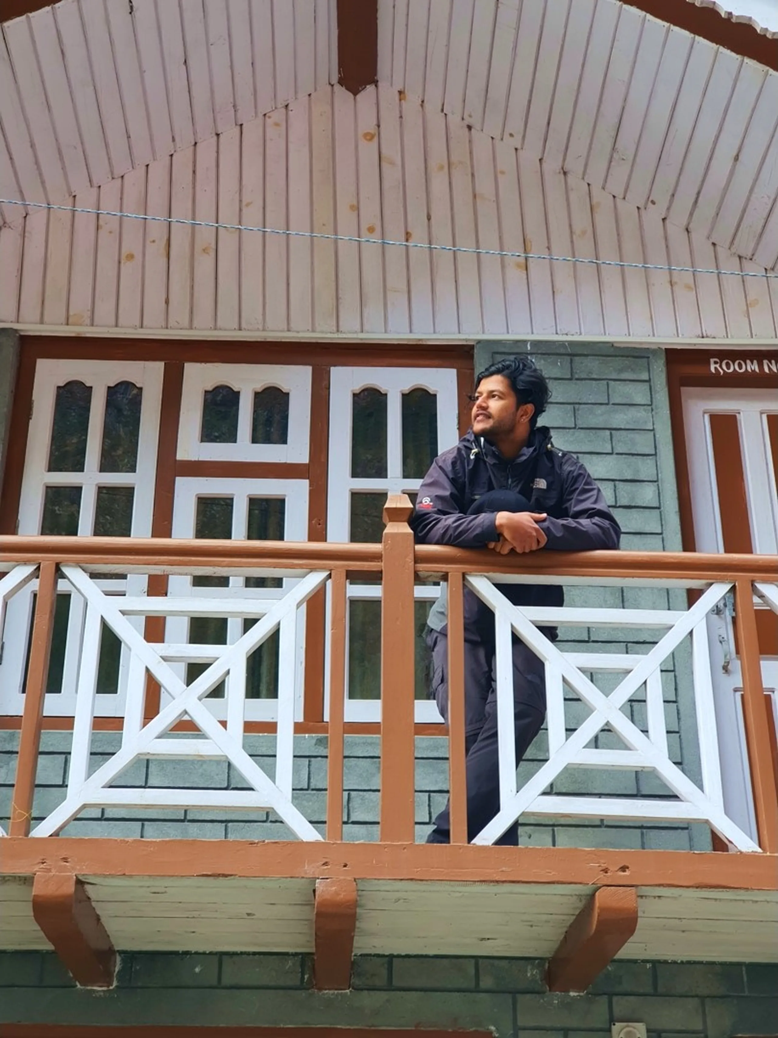 A pensive young male Nepali trekker leans over the wooden railing of a second-floor balcony at a Himalayan guesthouse in Manang, Nepal - wearing a dark grey North Face trekking jacket, arms folded on the railing, gazing thoughtfully into the distance - the traditional Nepali lodge facade features grey brick walls, ornate white-trimmed windows with carved lattice patterns, and a curved corrugated metal roof - a 'Room No.' sign partially visible - a quietly atmospheric Nepal trekking lodge portrait capturing the contemplative moments between trekking stages on the Annapurna Circuit