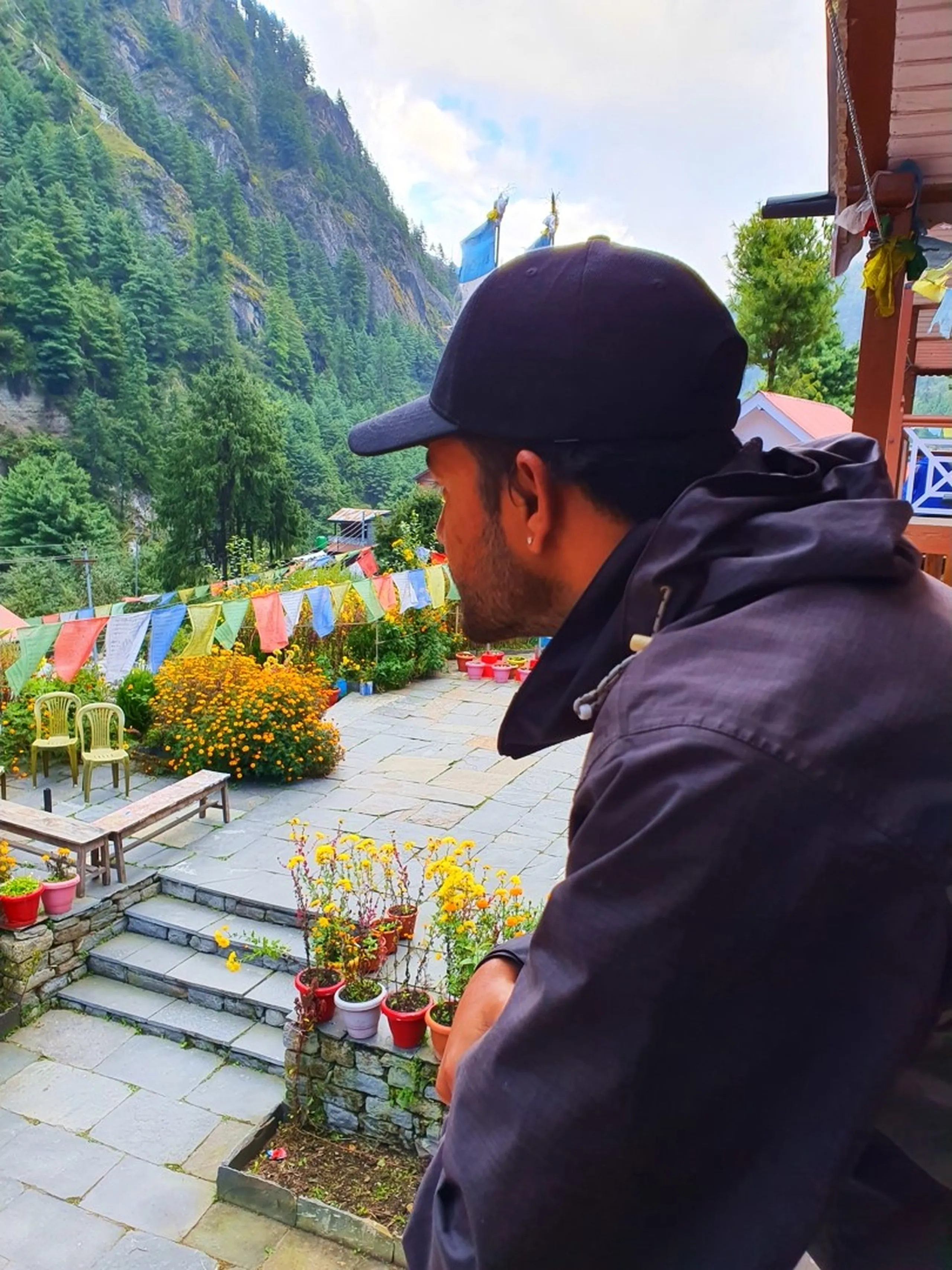 A rear-view portrait of a young male trekker in a black hoodie and black cap sitting on the edge of a traditional Nepali teahouse terrace in Chame, Manang, gazing over a beautifully landscaped stone-paved courtyard garden - rows of colorful Buddhist Tibetan prayer flags in red, yellow, blue, green, and white are strung diagonally across the garden - vibrant orange and yellow marigold flowers bloom in red terracotta pots arranged along stone steps - yellow plastic chairs and a wooden bench sit in the garden - dense pine-forested Himalayan cliffs rise steeply in the background under a cloudy sky - a serene and culturally rich Nepal trekking lodge garden scene on the Annapurna Circuit, Chame, Manang