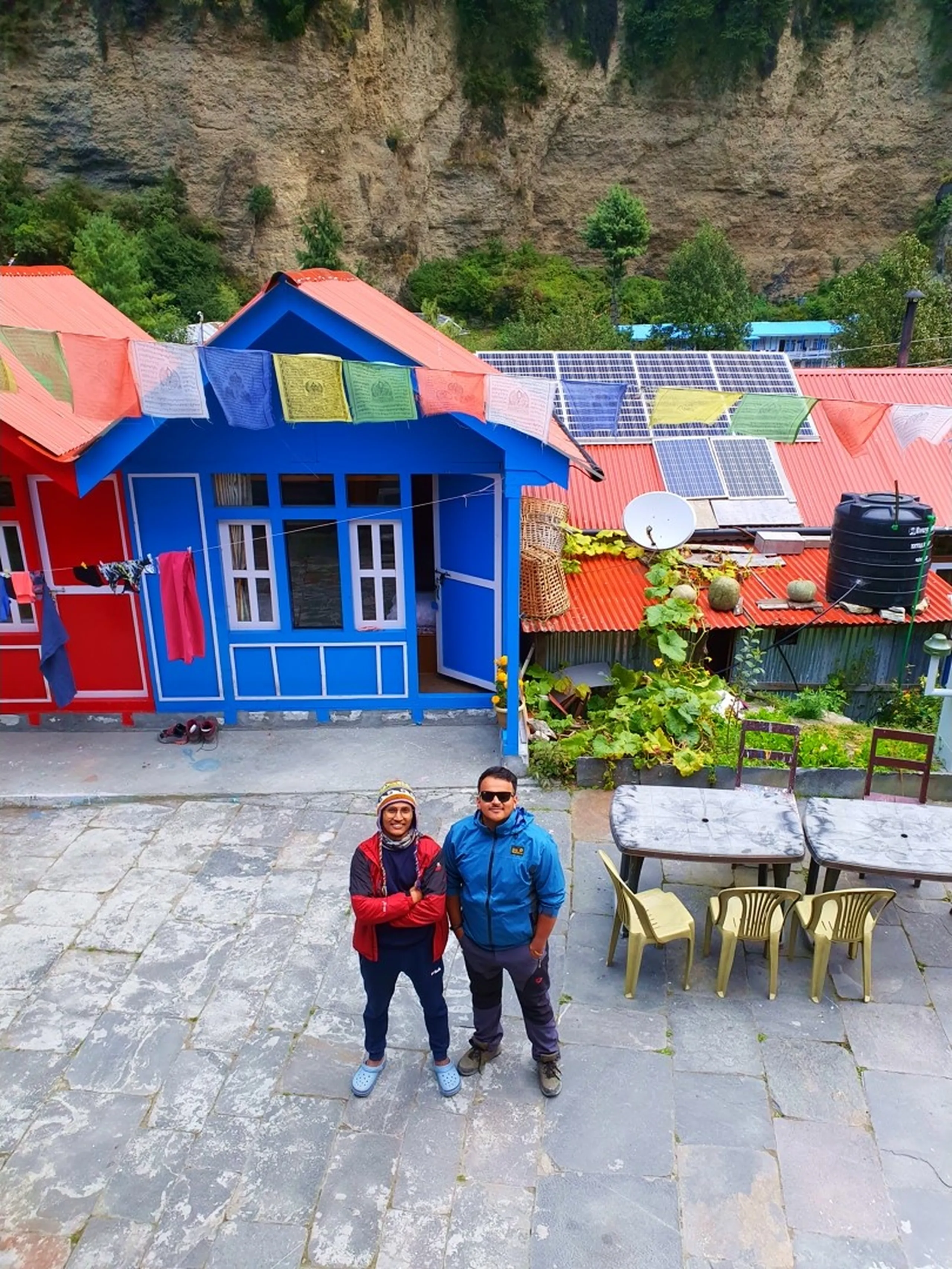 A bird's-eye view looking down on two young male Nepali trekkers standing together in the stone-paved courtyard of a vibrantly painted trekking lodge in Chame, Manang, Nepal - one wearing a traditional Nepali wool hat, red jacket with arms crossed, the other in a blue Jack Wolfskin jacket and sunglasses - behind them a bold cobalt blue and orange-red painted wooden guesthouse with colorful Tibetan Buddhist prayer flags strung across the rooftop - solar panels, a water tank, a satellite dish, and a vegetable garden visible on the adjacent orange corrugated roof - rough cliff rock face rising directly behind - an authentic Nepal mountain village guesthouse scene on the Annapurna Circuit trekking route