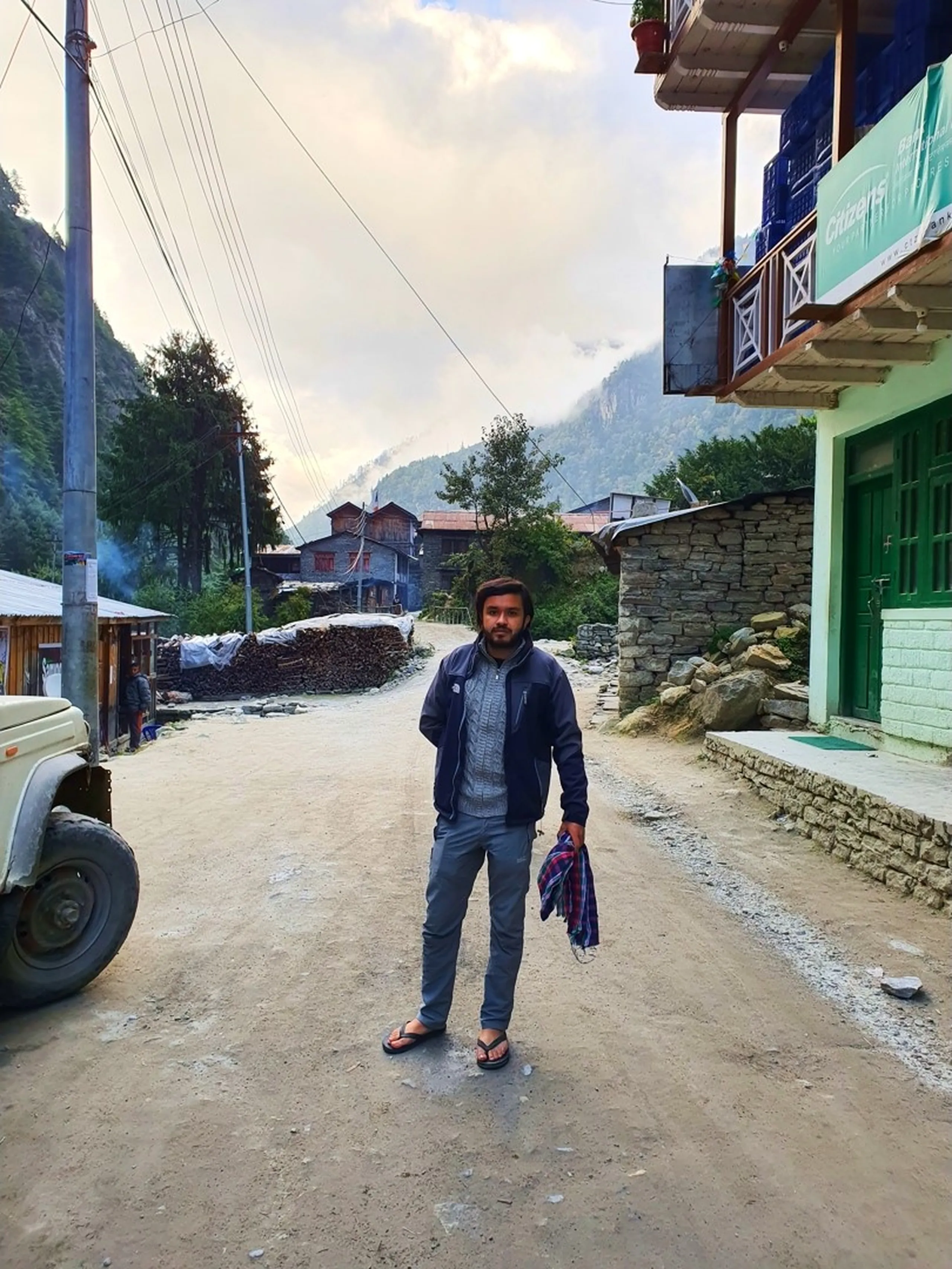 A young male trekker stands alone in the center of a quiet early-morning mountain village street in Chame, Manang, Nepal - wearing a dark navy North Face zip jacket, grey trousers, flip flops, and carrying a folded flannel shirt - traditional stone-built teahouse buildings line both sides of the dusty road, a Citizen's Bank sign and a green building visible to the right, a 4WD jeep parked to the left - firewood stacked in the middle distance, smoke drifting from a chimney - misty cloud-capped mountains visible at the end of the village lane - a candid and atmospheric Nepal Himalayan village street photograph on the Annapurna Circuit trekking route to Manang