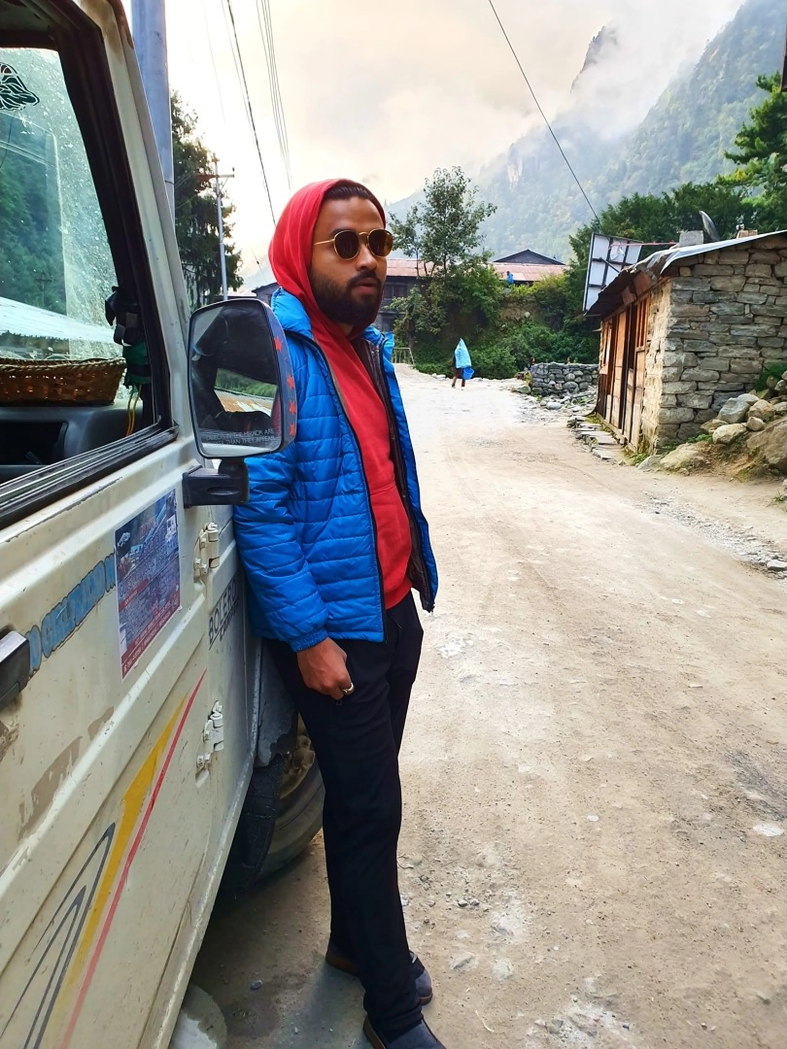 A stylish young male trekker leans coolly against the side door of a white Mahindra Bolero jeep in a misty mountain village lane in Chame, Manang, Nepal - wearing a bright red hoodie under a vivid blue puffer jacket, black trekking trousers, dark shoes, and round gold-framed sunglasses - looking away from the camera with a composed expression - traditional stone Himalayan lodge buildings and a misty valley with pine forest and grey rock cliffs visible behind - a moody and cinematic Nepal trekking portrait in a Manang village, Annapurna Circuit, capturing the style and spirit of adventure travel in Nepal