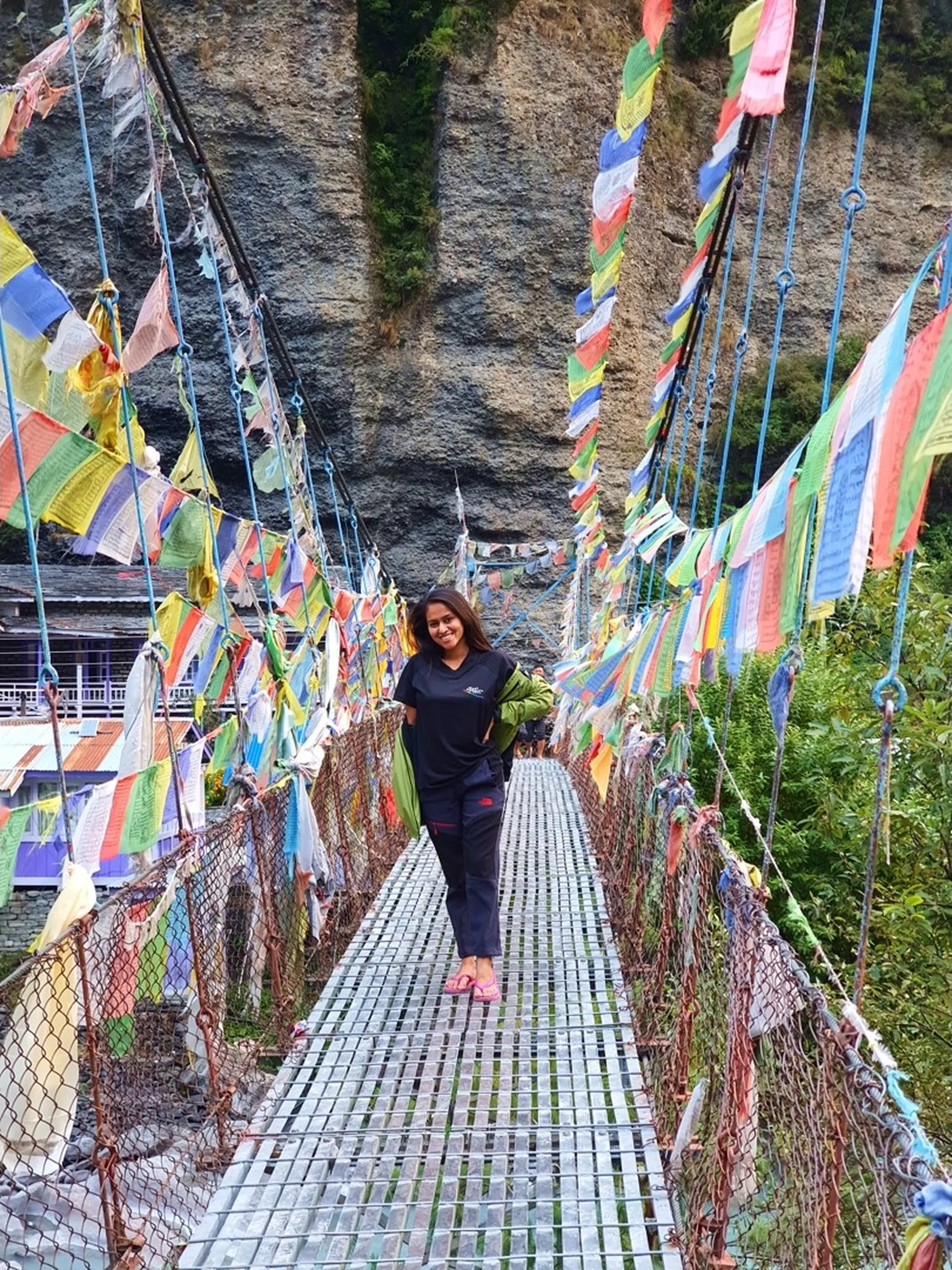 A young Nepali woman stands smiling in the center of a colorful suspension footbridge in Manang, Nepal - completely adorned with hundreds of vibrant Tibetan Buddhist prayer flags in red, yellow, white, blue, and green fluttering from cables on both sides - wearing a dark navy Great Himalayan Trail tee, black trekking trousers, and pink flip flops - a towering bare sandstone cliff face fills the entire background - a small Nepali building visible at the far end of the bridge - a stunning and culturally layered Nepal trekking bridge photograph, one of the most photogenic prayer flag bridges on the Annapurna Circuit, ideal for Nepal adventure travel and Buddhist culture blog content
