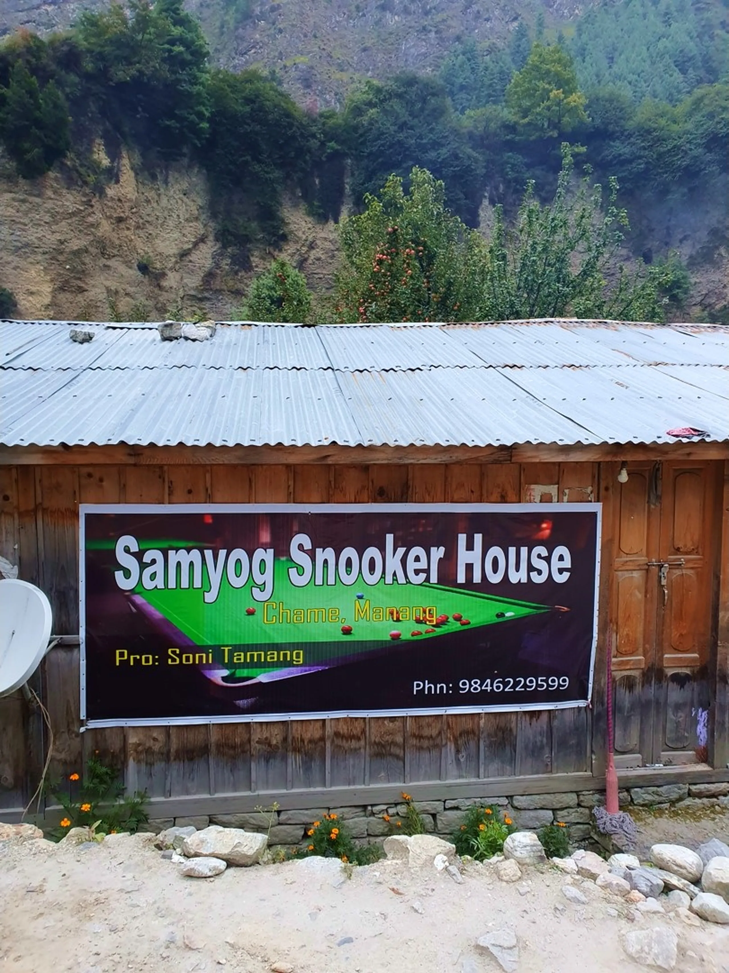A wooden-clad roadside building in Chame, Manang, Nepal with a corrugated metal roof displaying a large green and black banner reading 'Samyog Snooker House - Chame, Manang - Pro: Soni Tamang - Phn: 9846229599' - a satellite dish mounted to the left, marigold flowers growing at the base, a fruit tree with red apples visible on the rooftop - rocky Himalayan hillside rising steeply in the background under grey overcast skies - a surprisingly charming slice of local village life in a remote Himalayan trekking town, a quirky and memorable stop on the Annapurna Circuit to Manang, Nepal - great for Nepal offbeat travel and hidden gems blog content