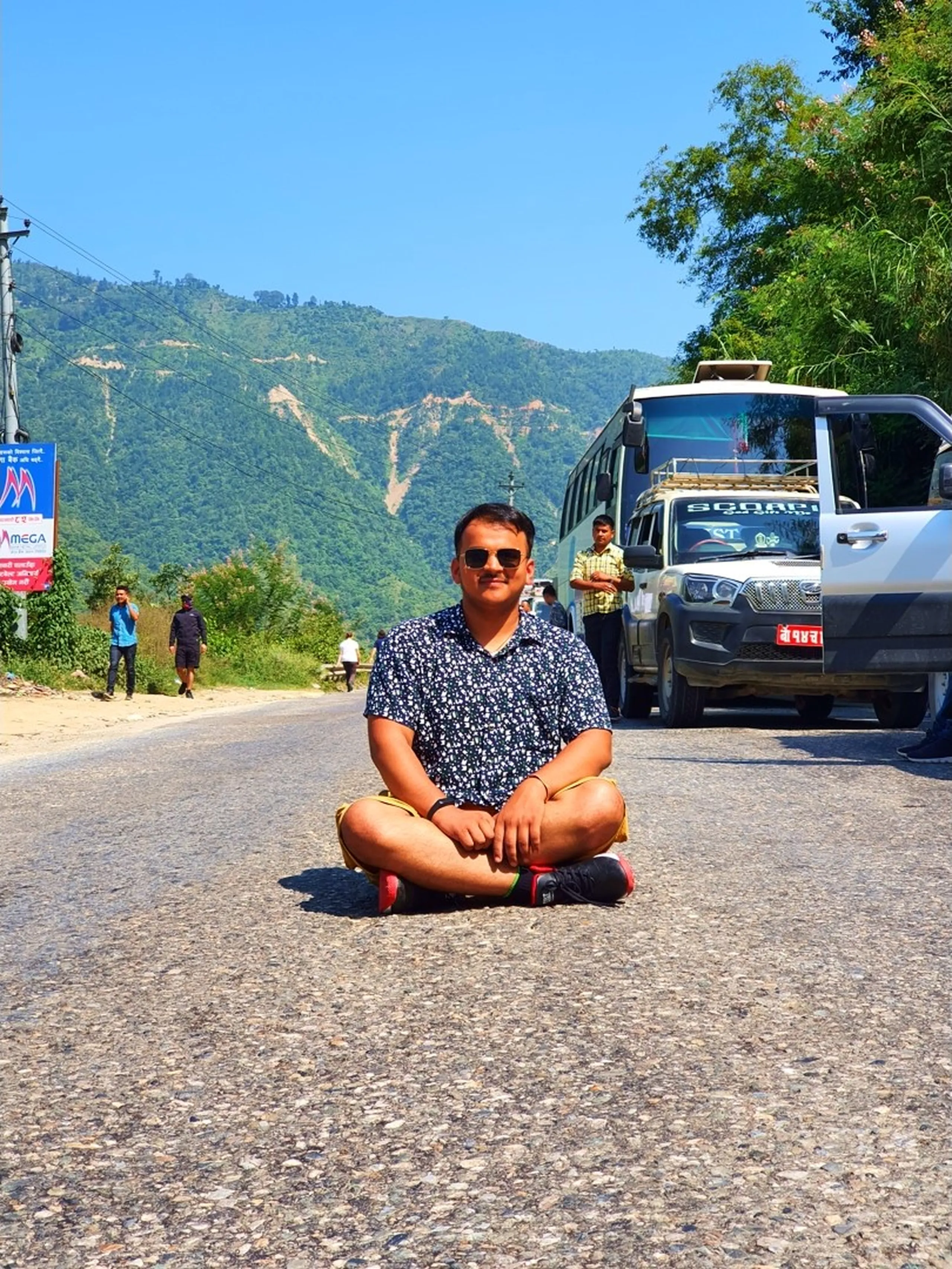 A young male traveler sits confidently cross-legged in the center of a quiet mountain highway in Nepal, wearing a patterned navy shirt, yellow shorts, and black sneakers, posing casually with traffic backed up behind him - a tourist bus and a white Scorpio SUV visible, green forested hills and a Mega Bank signboard in the background under brilliant blue skies - a bold and fun Nepal road trip photo perfect for travel blogs covering Kathmandu to Manang trekking routes