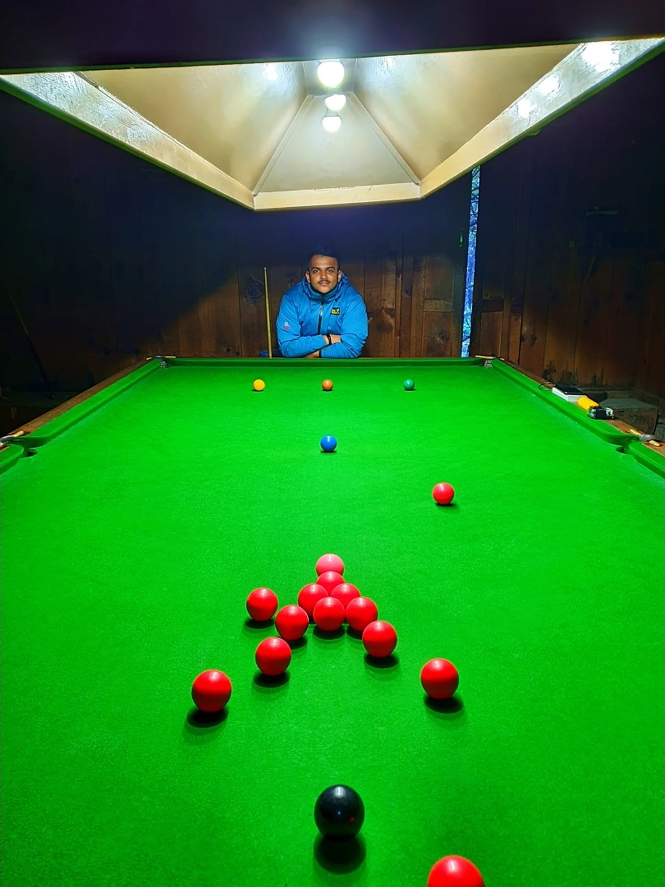 A wide-angle shot of a young male Nepali trekker leaning forward over a full-size green-baize snooker table inside a dimly lit wooden snooker hall in Chame, Manang, Nepal - arms crossed, looking directly at the camera - red balls racked in a triangle formation in the foreground, with blue, orange, green, and black balls scattered across the table - a pyramid of ceiling lights illuminates the table from above - wooden panel walls surround the dark interior - a uniquely unexpected Nepal mountain travel photo capturing a snooker hall found in one of the most remote villages on the Annapurna Circuit, perfect for Nepal offbeat travel blog and hidden gems of Manang content