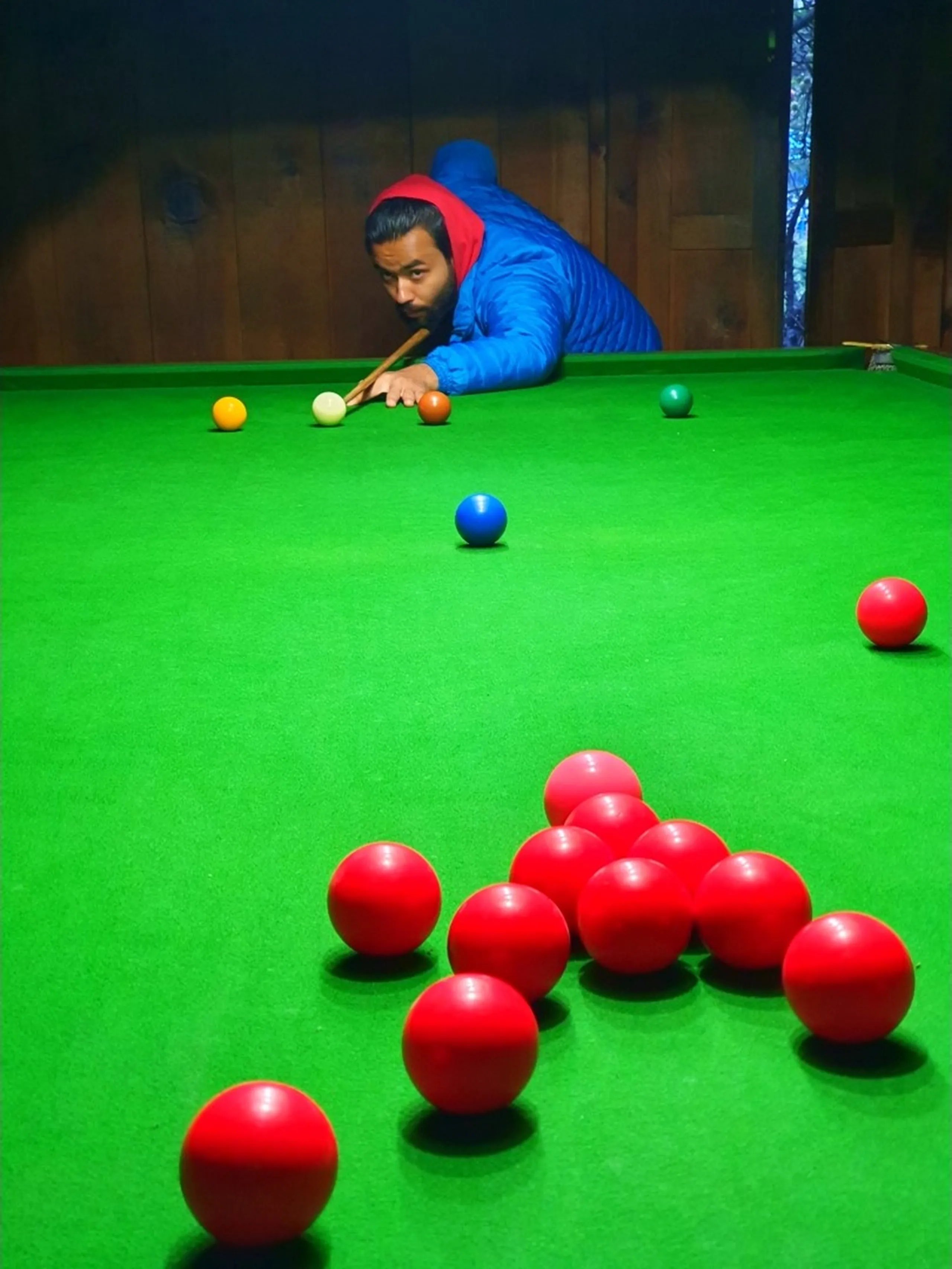 A young male Nepali trekker leans intently over a bright green snooker table in a dimly lit wooden hall in Chame, Manang, Nepal - wearing a red hoodie under a blue puffer jacket - cue in hand, carefully lining up a shot on the cue ball surrounded by red, orange, yellow, blue, and green balls - his focused expression and playing stance captured mid-game - a surprisingly fun and unexpected Nepal mountain travel moment, playing snooker in one of the most remote teahouse villages on the Annapurna Circuit, showcasing the unique and offbeat experiences of trekking through Manang, Nepal