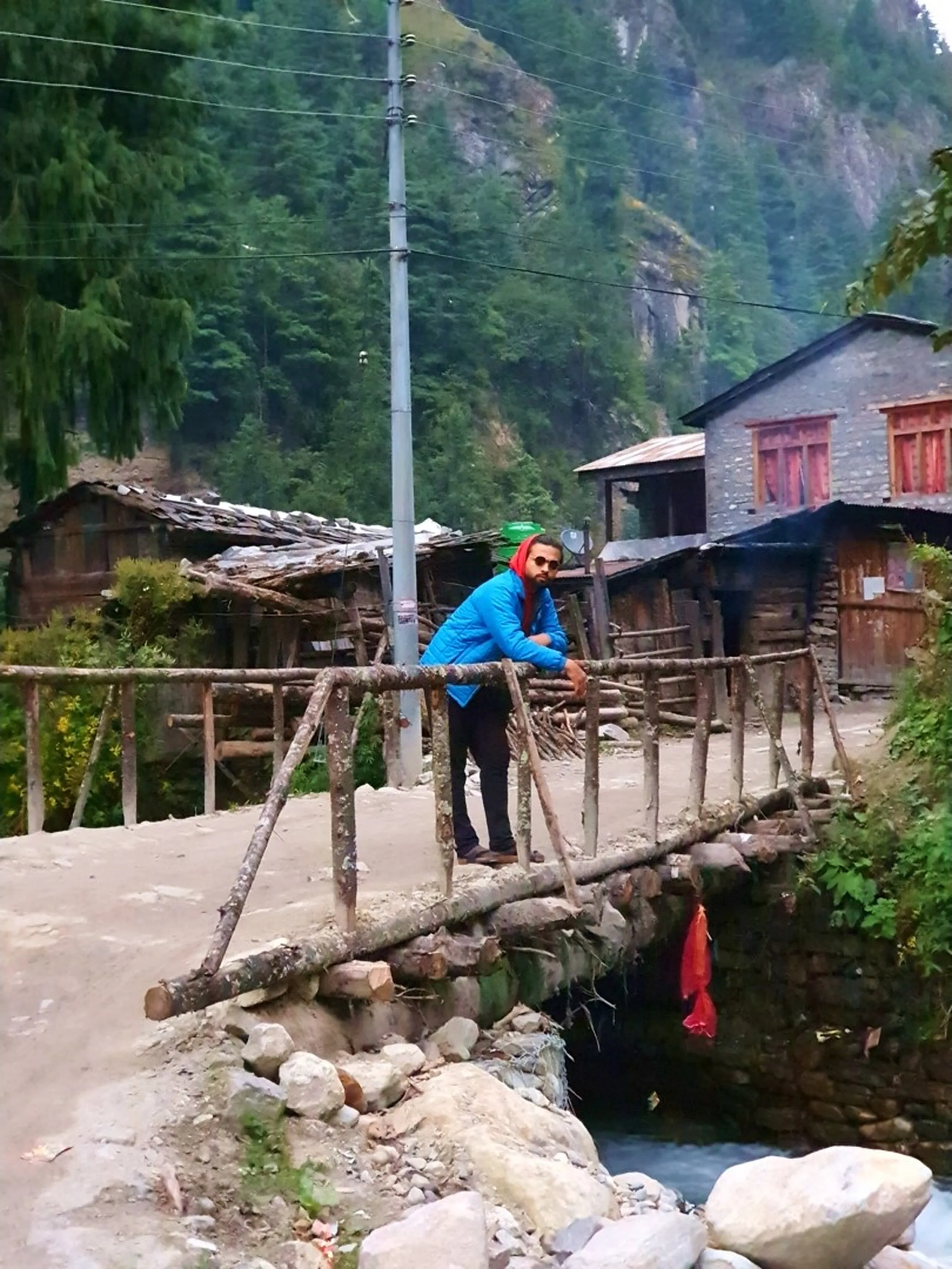 A young male Nepali trekker in a bright red hoodie and blue puffer jacket stands leaning on the railing of a rough-hewn log-and-stone footbridge over a small rushing mountain stream in a remote village in Chame, Manang, Nepal - looking directly at the camera - traditional stone Himalayan teahouse buildings with wooden roofs visible behind him, dense pine forest climbing steep cliffs above - a red cloth offering tied to the bridge railing - grey overcast sky - a characterful and authentic Nepal mountain village portrait on the Annapurna Circuit, perfectly capturing the rustic charm of trekking through remote Himalayan settlements