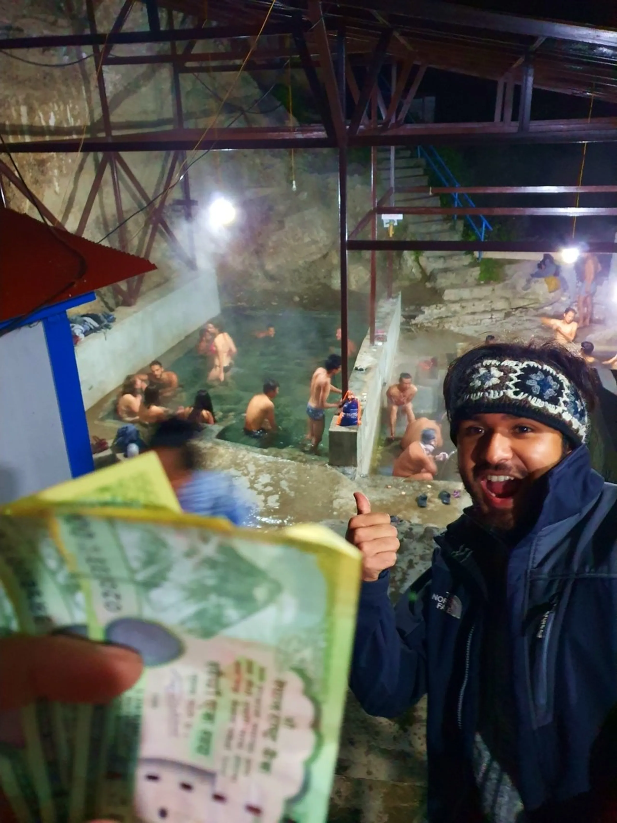 A joyful young male Nepali trekker in a black North Face jacket and black-white patterned wool headband gives an enthusiastic thumbs-up while grinning widely at the camera - he holds a fan of Nepali rupee banknotes in his other hand - behind him, looking down from an elevated vantage point, is a large natural hot spring bathing pool full of locals and trekkers soaking in the steaming thermal water - the pool is covered by an open industrial roof structure with bright overhead lights and surrounded by stone walls - a thrilling and energetic Nepal natural hot spring experience photograph, the famous Tatopani hot spring on the Annapurna Circuit, one of Nepal's most beloved trekking rest stops