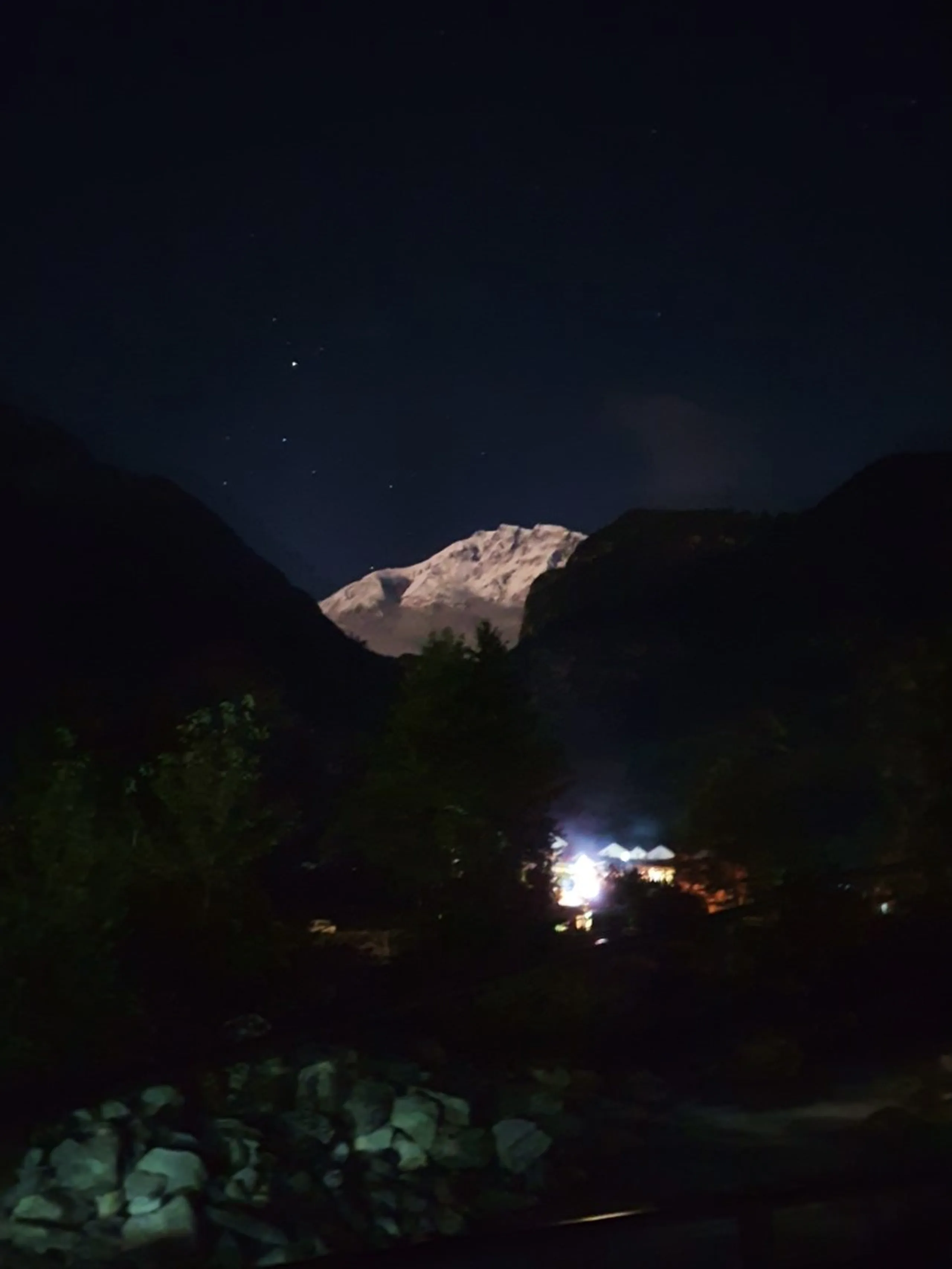 A dramatic low-light night photograph of a snow-capped Himalayan peak glowing in moonlight between two dark silhouetted mountain ridges in Manang, Nepal - the white glacier-covered summit luminous against the deep navy blue starry sky - a cluster of warm glowing teahouse lights visible at the base of the valley - silhouetted pine trees in the mid-ground - a deeply atmospheric and romantic Nepal Himalayan night landscape photograph, showcasing the majestic Annapurna mountain range at night as seen from a Manang trekking lodge, ideal for Nepal night photography and Annapurna Circuit stargazing blog content