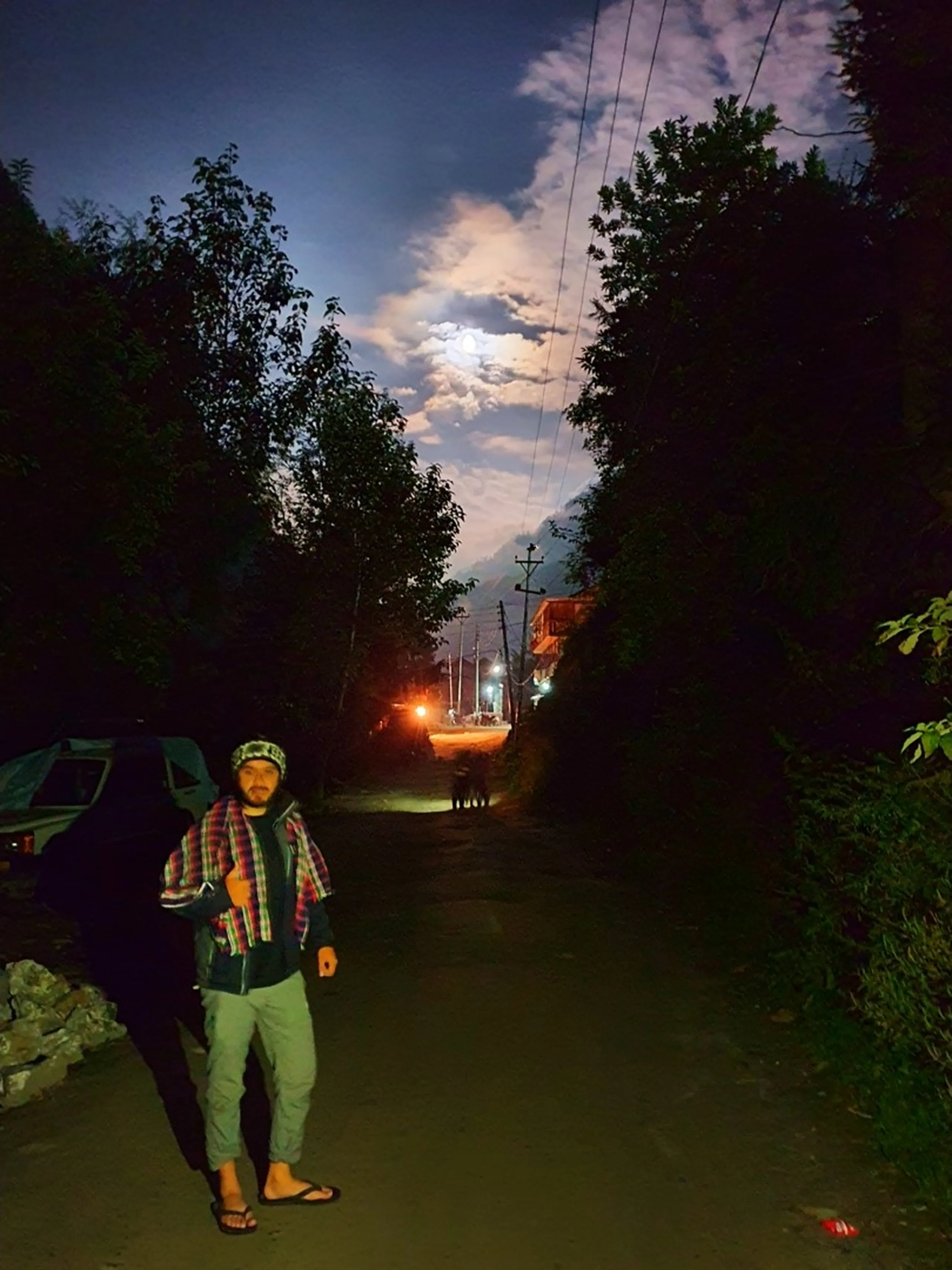 A young male Nepali trekker stands on a dark village lane at night in Chame, Manang, Nepal - wearing a plaid flannel shirt over a dark fleece jacket, grey trekking trousers, flip flops, and a black-and-white Nepali wool topi hat - posing casually on the dirt road flanked by tall dark trees - a warm amber street light glows in the distance and a full moon partially covered by dramatic backlit clouds illuminates the sky above - a dog silhouette visible in the mid-ground near the light - a moody and cinematic Nepal mountain village night portrait on the Annapurna Circuit, capturing the peaceful and atmospheric evenings in Manang