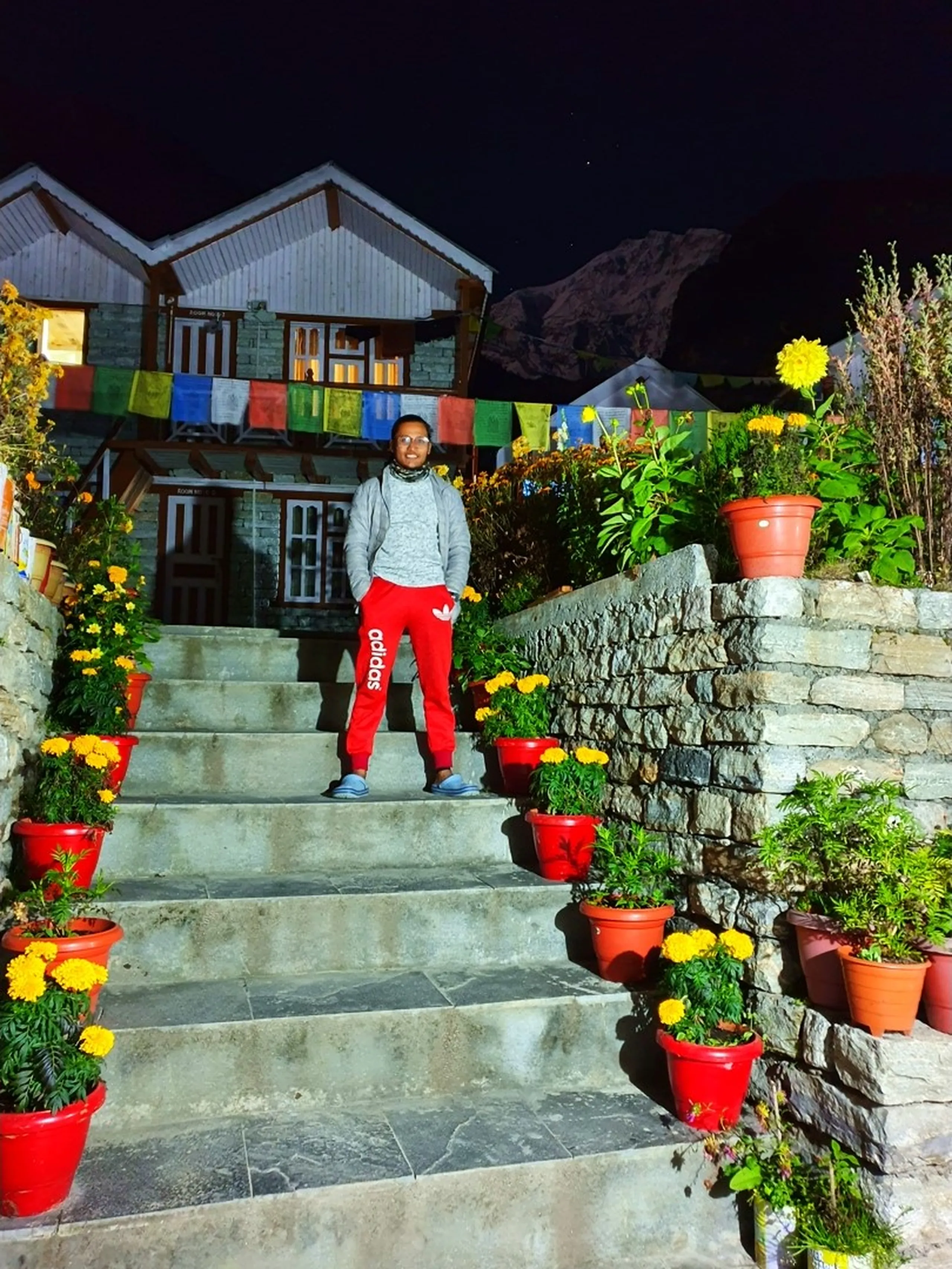 A young male trekker stands on stone steps outside a traditional Nepali mountain lodge at night in Manang, Nepal - wearing bright red Adidas track pants, a grey sweatshirt, glasses, and blue sandals - surrounded by vibrant red terracotta pots bursting with yellow marigold flowers lining every step - colorful Tibetan Buddhist prayer flags in red, blue, yellow, green, and white are strung across the front of the guesthouse behind him, whose warm amber windows glow in the darkness - a jagged dark mountain silhouette rises behind the building against the black night sky - a vivid and culturally textured Nepal trekking lodge night portrait, showcasing the beautiful flower-lined guesthouses of Manang on the Annapurna Circuit