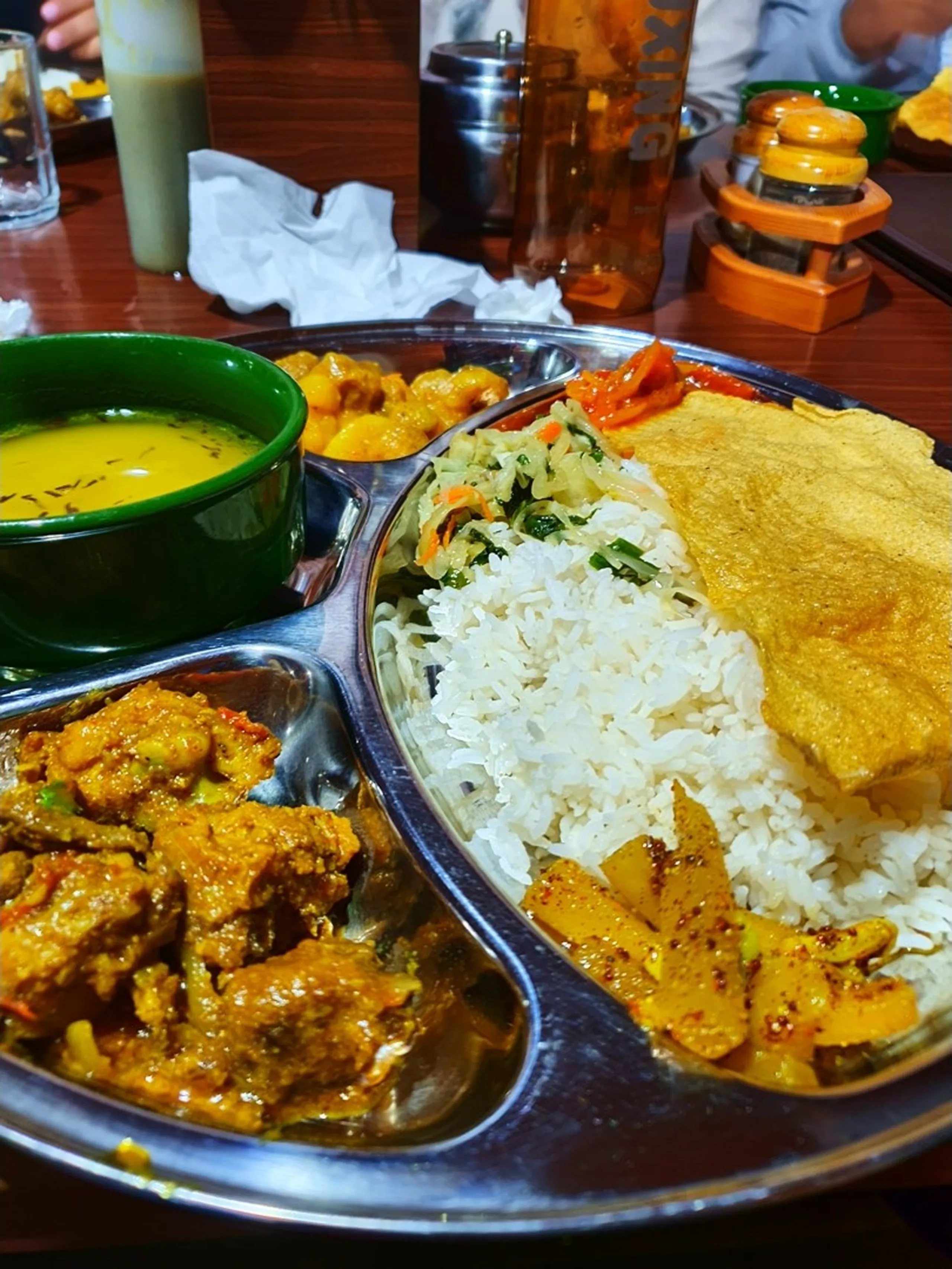 A close-up overhead shot of a hearty Nepali trekking dinner served on a stainless steel divided thali plate on a wooden restaurant table in Manang, Nepal - featuring a generous mound of steamed white rice topped with a large golden egg omelette, spiced potato and pumpkin curry, a fresh vegetable coleslaw salad with carrots, a rich chicken curry with chunky pieces in brown gravy, a side dish of cauliflower curry, a small tomato achar, and a green ceramic bowl filled with bright yellow lentil dal - a trekking water bottle and salt-and-pepper set visible in the background - authentic high-altitude Nepali comfort food photography from the Annapurna Circuit, ideal for Nepal trekking food blog and dal bhat culture content