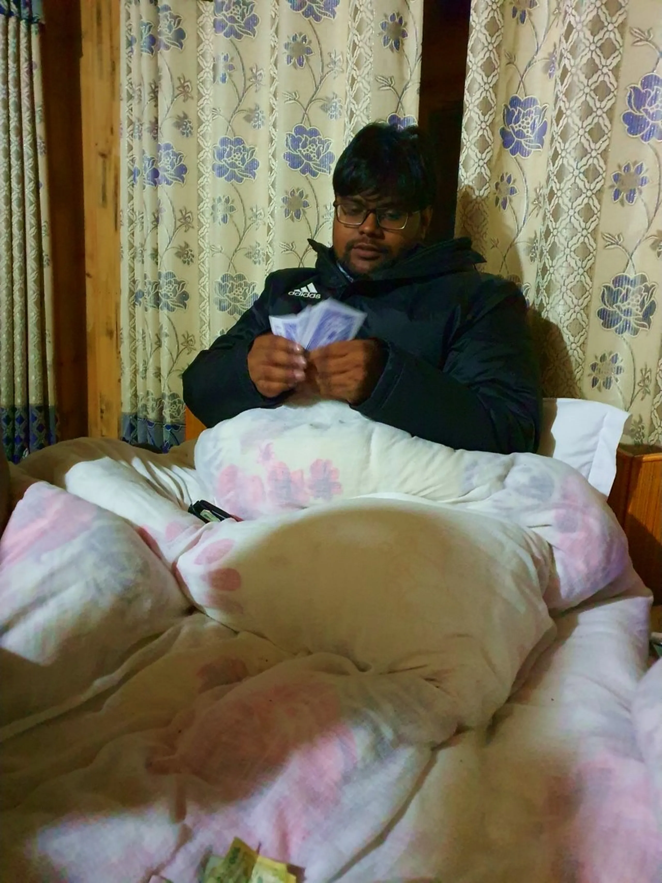 A candid and humorous Nepal trekking lodge moment - a heavyset young male trekker wearing glasses and a black Adidas jacket sits upright in a cozy guesthouse bed in Manang, Nepal, surrounded by thick floral-patterned pillows and blankets pulled up to his waist - counting or shuffling a hand of playing cards with intense concentration - wooden walls, a floral curtain behind him, and another set of cards on the bed visible - a warm and relatable Nepal teahouse night scene, capturing the fun indoor pastimes trekkers enjoy after a long day on the Annapurna Circuit trail, perfect for Nepal trekking lifestyle blog content