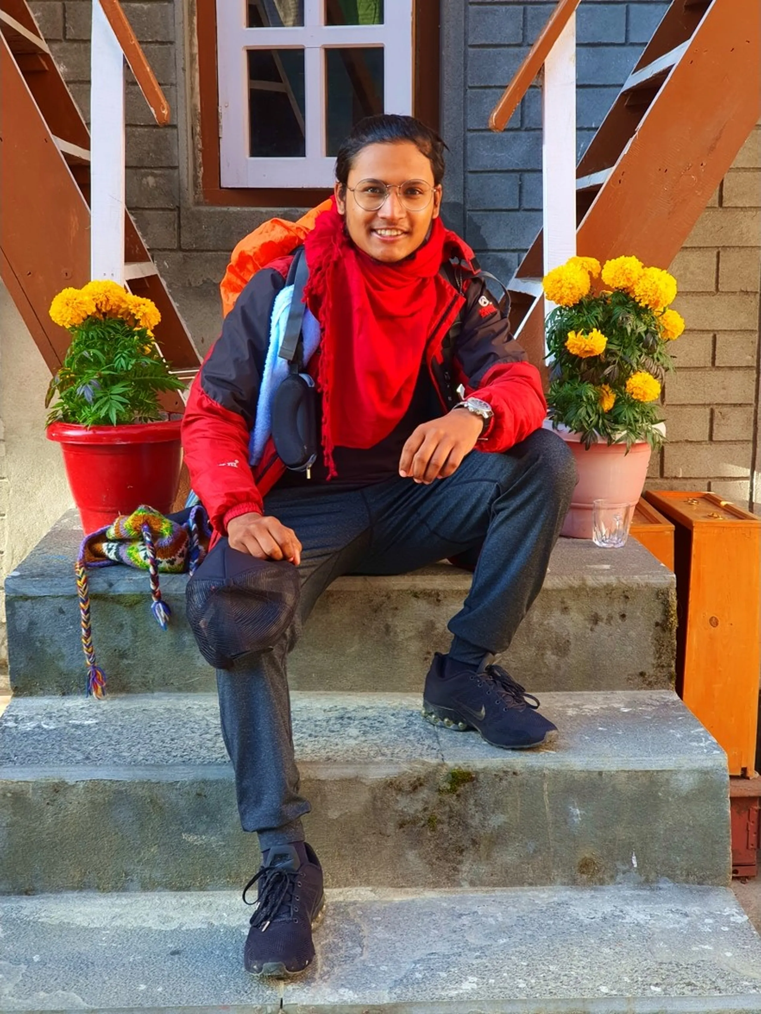 A cheerful young male trekker sits on the stone entrance steps of a traditional Nepali guesthouse in Manang, Nepal, fully geared up with a large orange trekking backpack - wearing a red jacket layered over a red scarf, grey trekking trousers, Nike running shoes, and round wire-framed glasses - a colorful Nepali wool friendship bracelet resting on the step beside him, vibrant yellow marigold flowers in red pots flanking the steps - smiling broadly and ready for the day's trek - a warm and energetic Nepal trekking departure portrait at a Manang lodge, capturing the morning excitement and gear-ready spirit of trekkers on the Annapurna Circuit heading towards Ice Lake