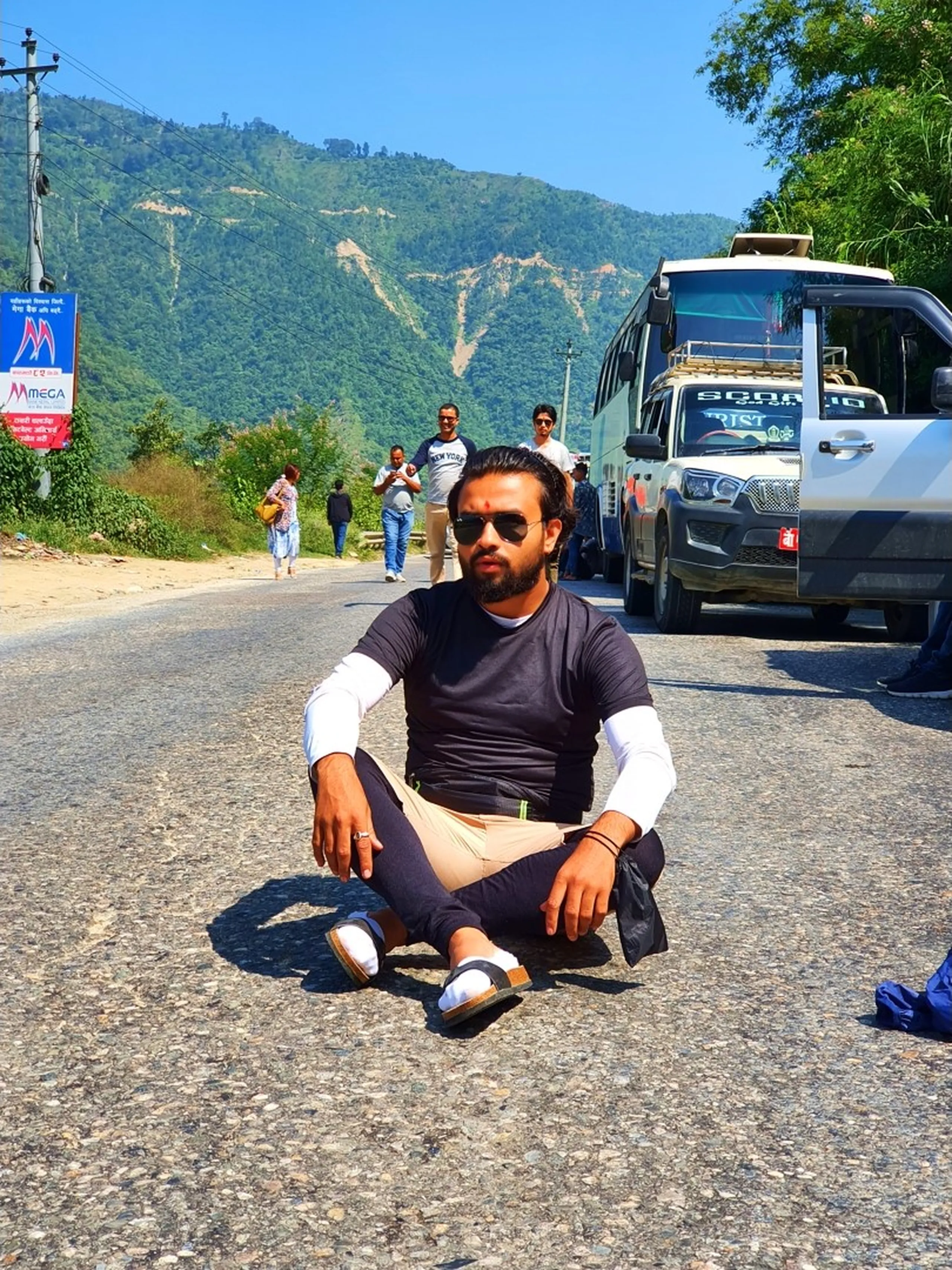 A young male Nepali trekker sits relaxed on a rural mountain road in Nepal, wearing a black long-sleeve layered shirt, beige pants, sandals, and dark sunglasses, with a tika dot on his forehead, hands resting casually on his knees - a tourist bus, white Scorpio, other travelers, and lush green Himalayan hills fill the background under a crisp blue sky - a spontaneous travel moment during a Nepal Himalaya road trip, Kathmandu to Manang trekking