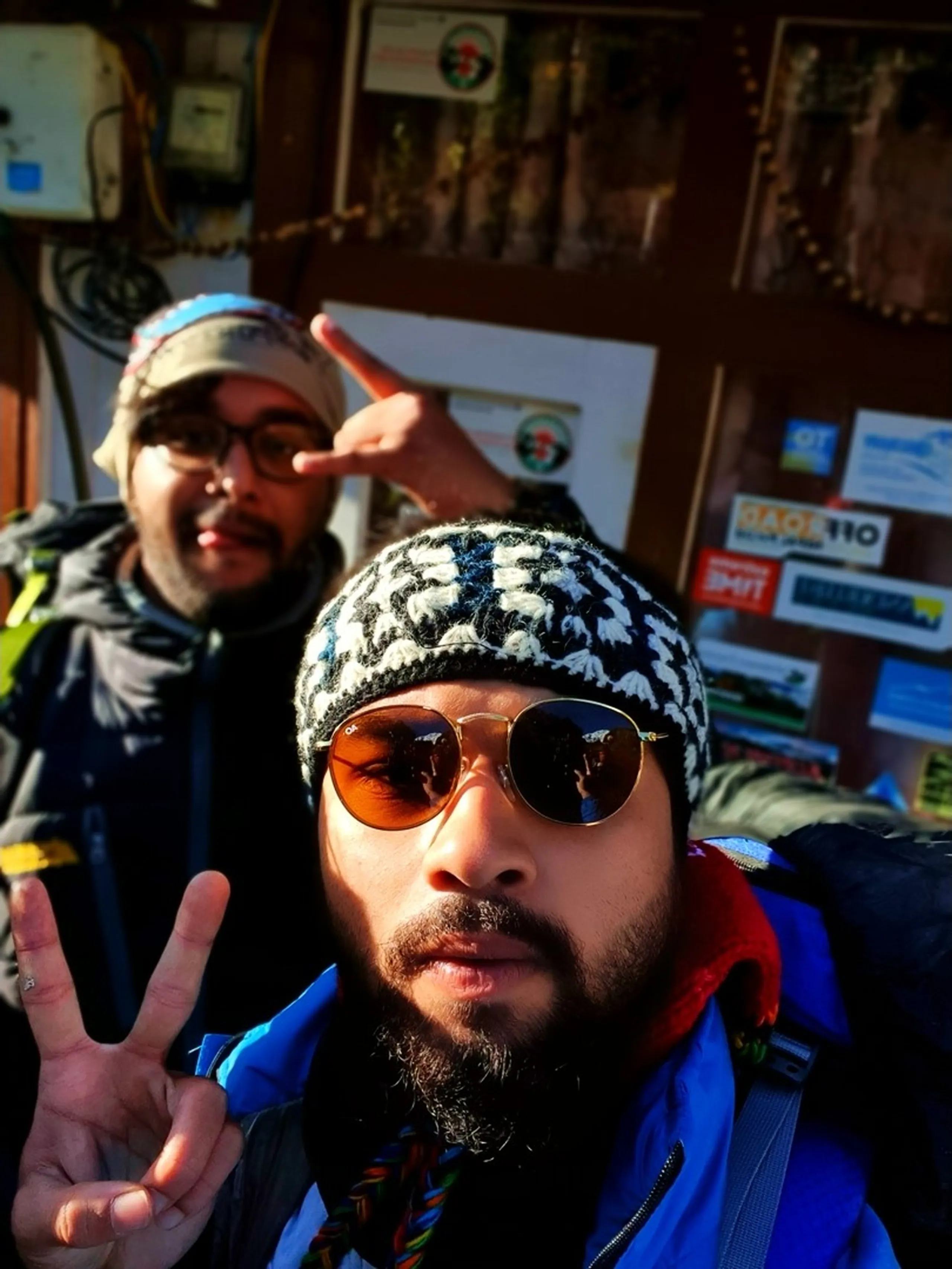 A close-up selfie of two young male Nepali trekkers fully geared up inside a teahouse checkpoint in Manang, Nepal - the one in front wearing a black-and-white traditional Nepali wool topi hat, round copper-framed aviator sunglasses, a blue puffer jacket, and a red scarf, flashing a peace sign - the one behind wearing glasses, a grey wool hat, and a dark jacket, also throwing a peace sign and grinning mischievously - trekking permit and sticker-covered wooden notice boards visible in the background - bright morning sunlight illuminating their faces - an energetic and joyful Nepal trekking checkpoint departure selfie on the Annapurna Circuit, capturing the excitement of setting off towards Ice Lake, Manang
