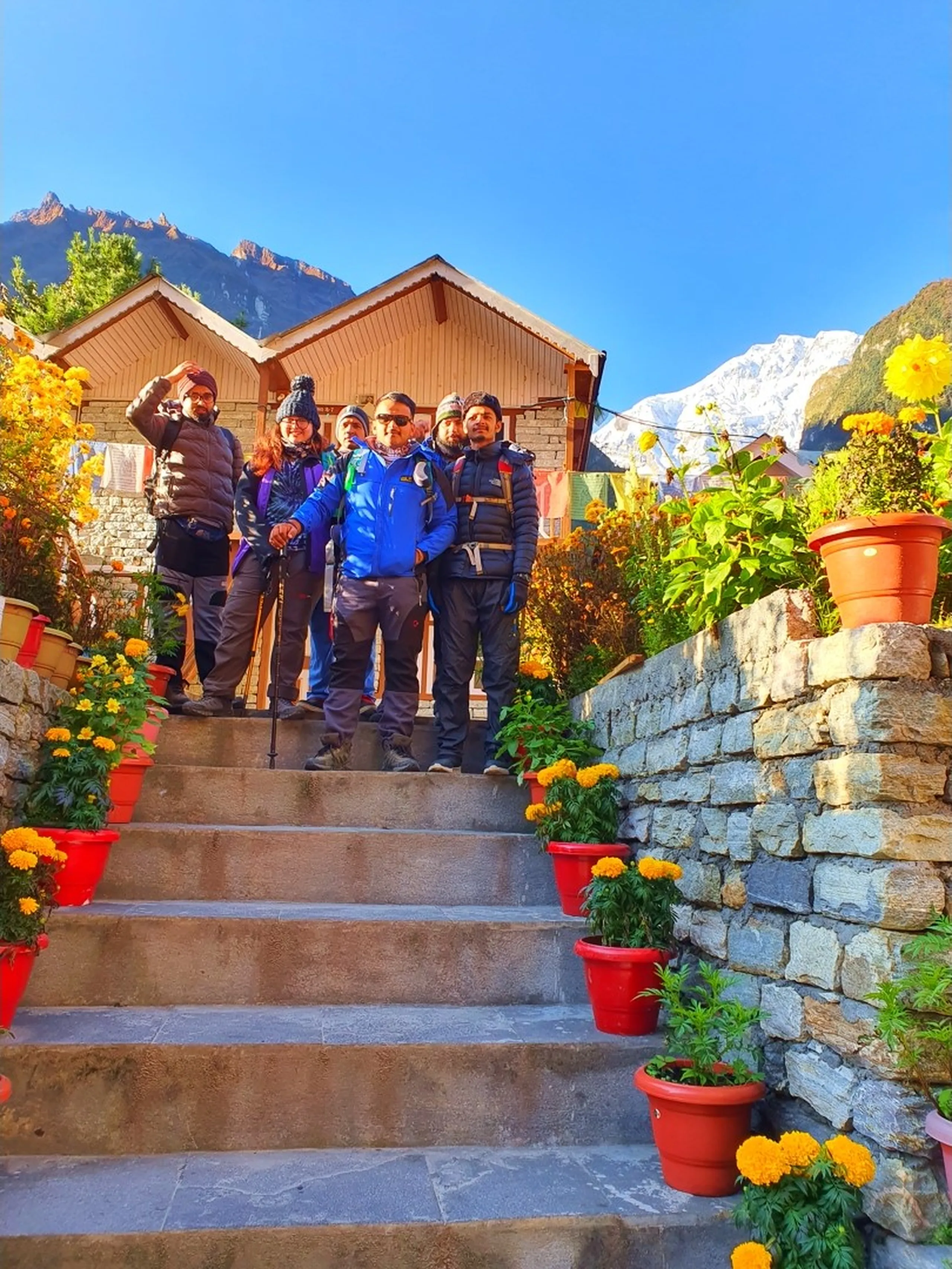 Five fully geared Nepali trekkers pose together on the marigold-lined stone entrance steps of a traditional mountain guesthouse in Manang, Nepal - wearing puffer jackets, wool hats, trekking pants, and loaded backpacks - one holding a trekking pole - vibrant yellow and orange marigold flowers in red terracotta pots line both sides of the steps - warm wooden lodge buildings behind them, and most spectacularly, a brilliant snow-capped Himalayan peak gleaming white against a deep blue sky - a classic and joyful Nepal trekking group departure photo, perfectly framing the Annapurna range at sunrise, ideal for Manang trekking itinerary and Annapurna Circuit group trek blog content