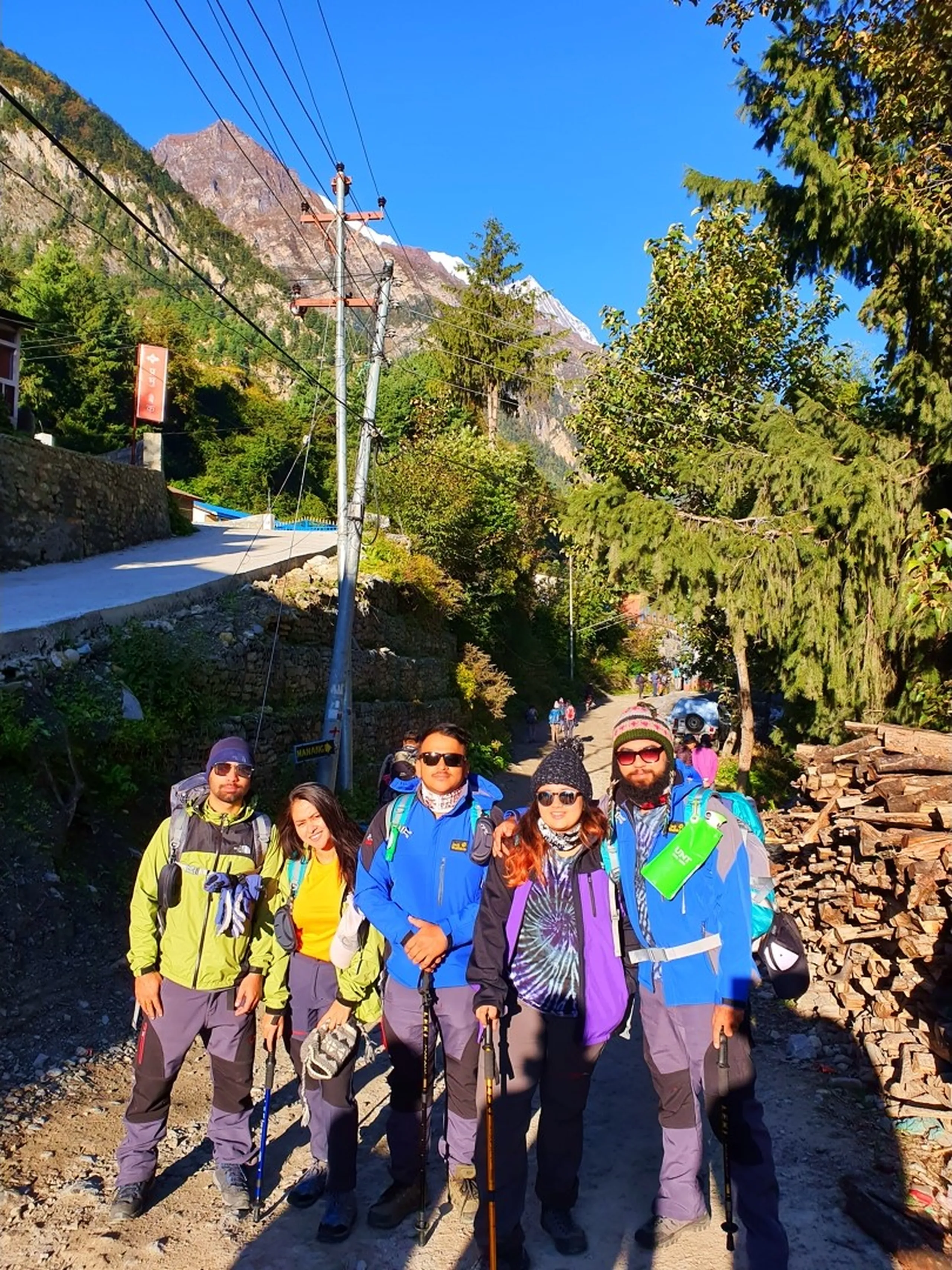 Five young Nepali trekkers fully loaded with backpacks and trekking poles pause together on a sunlit mountain dirt road in Chame, Manang, Nepal - wearing bright trekking jackets in neon green, yellow, and blue, with wool hats, sunglasses, and trekking trousers - smiling and posing confidently for the camera - a stacked firewood pile and traditional stone buildings visible behind them, pine trees lining the road, and a dramatic rocky Himalayan peak with a thin snow line rising against a brilliant deep blue sky - an energetic and vibrant Annapurna Circuit trekking trail group portrait, capturing the spirit of mountain adventure and camaraderie in Manang, Nepal