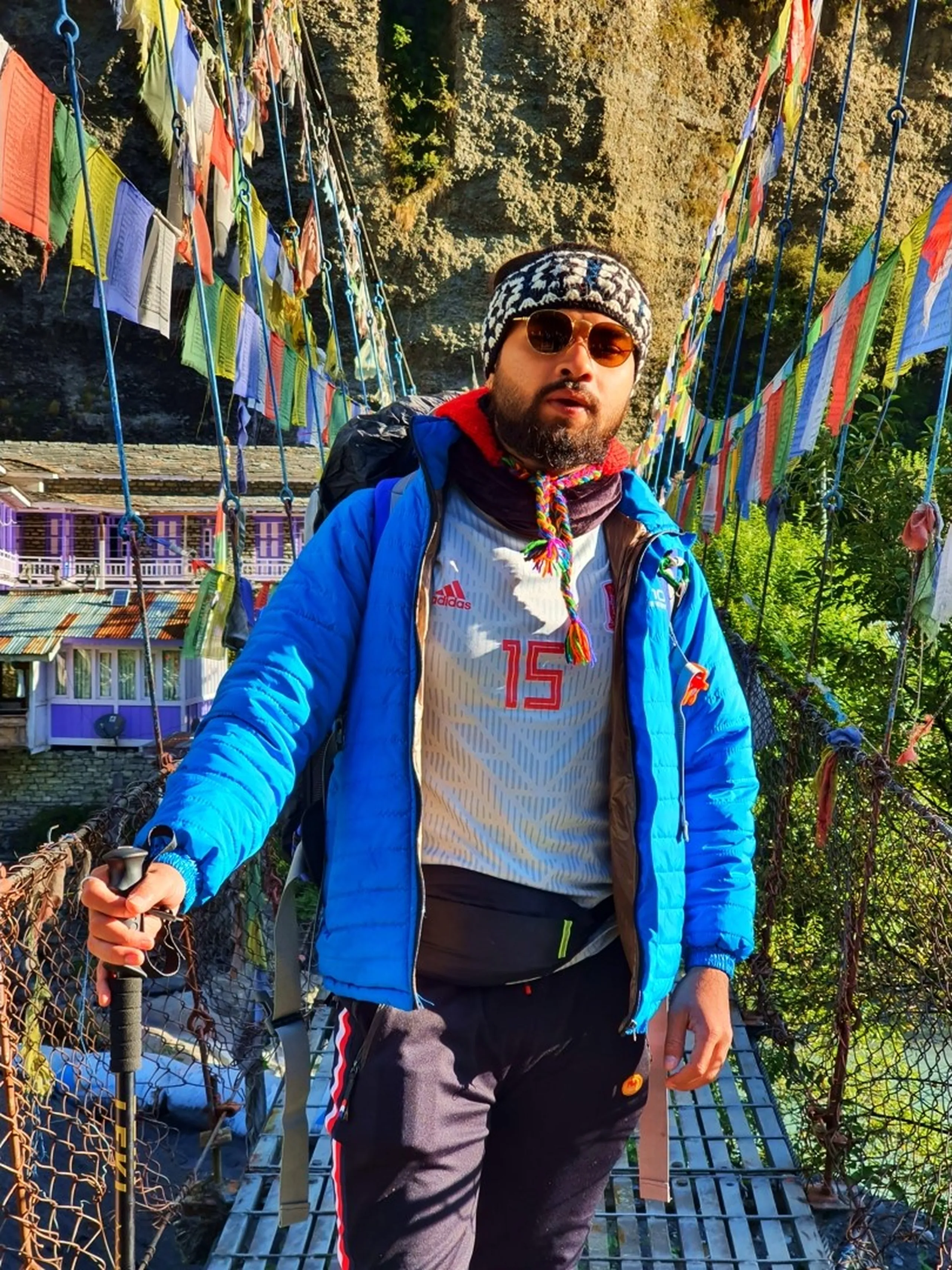 A close-up portrait of a young male Nepali trekker standing confidently on a colorful suspension footbridge in Manang, Nepal - wearing a blue puffer jacket open over a white and red Adidas number 15 football jersey, black track pants with red stripes, a black-and-white patterned Nepali wool headband, and copper round-framed aviator sunglasses - a colorful trekking scarf draped around his neck - holding a trekking pole in one hand, the other resting on the metal cable railing - hundreds of vibrant Tibetan Buddhist prayer flags in all colors flutter on both sides of the bridge - traditional Manang stone buildings and rocky cliffs visible behind - a bold and characterful Nepal adventure trekking portrait on the Annapurna Circuit prayer flag bridge