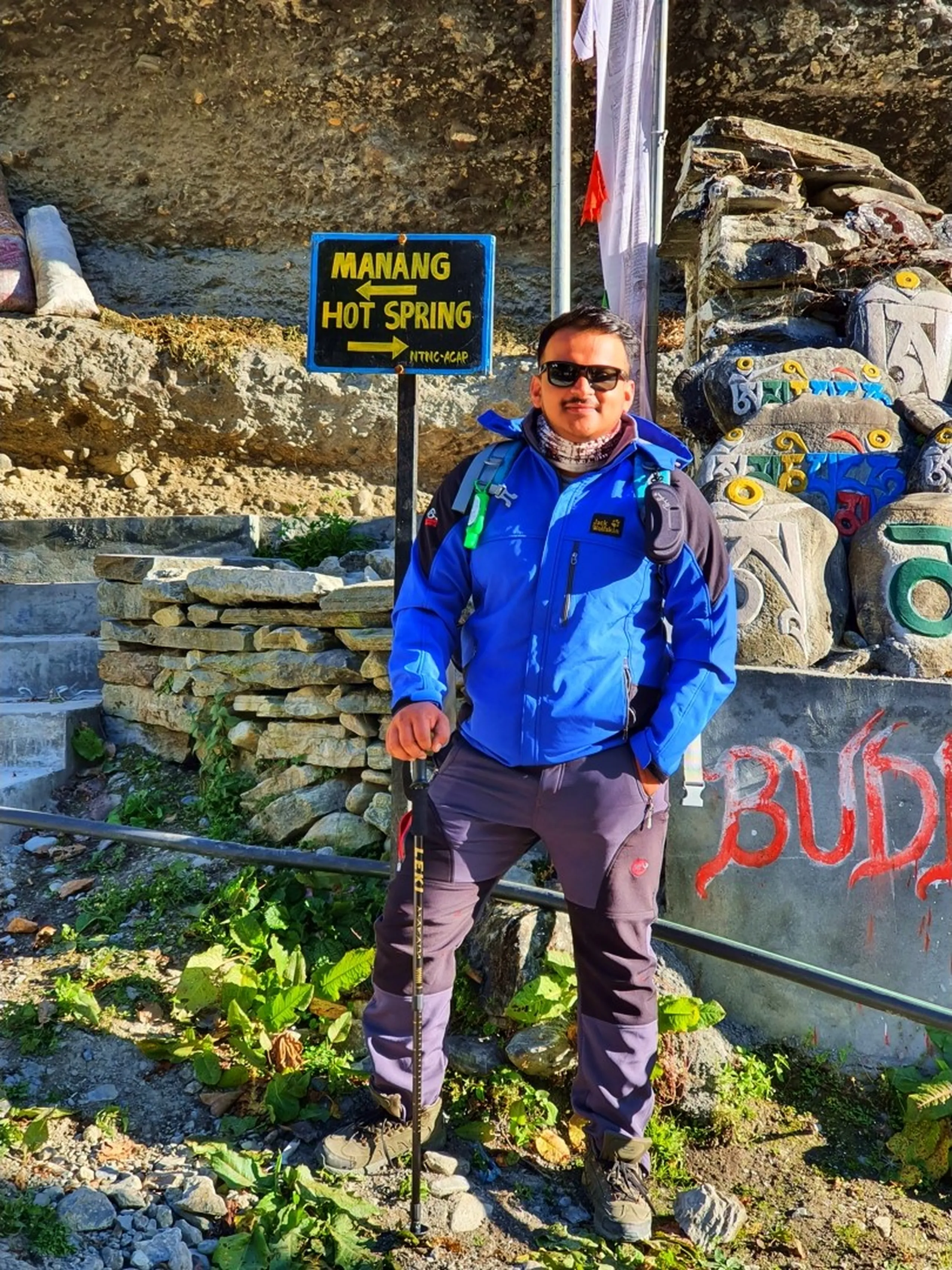 A male Nepali trekker stands confidently beside a black and yellow directional signpost reading 'Manang Hot Spring' with arrows pointing the way - wearing a bright blue Jack Wolfskin trekking jacket, dark grey Mammut trekking trousers, and hiking boots, holding a trekking pole - behind him a pile of colorfully painted Mani stones inscribed with Tibetan Buddhist mantras in green, blue, yellow, and red - a white prayer flag flutters from a pole above - 'BUDD' graffiti painted in red on a concrete wall - rocky cliffside backdrop - a culturally rich and practically useful Nepal trekking trail signpost photograph, highlighting the famous Manang Hot Spring on the Annapurna Conservation Area trekking route, ACAP