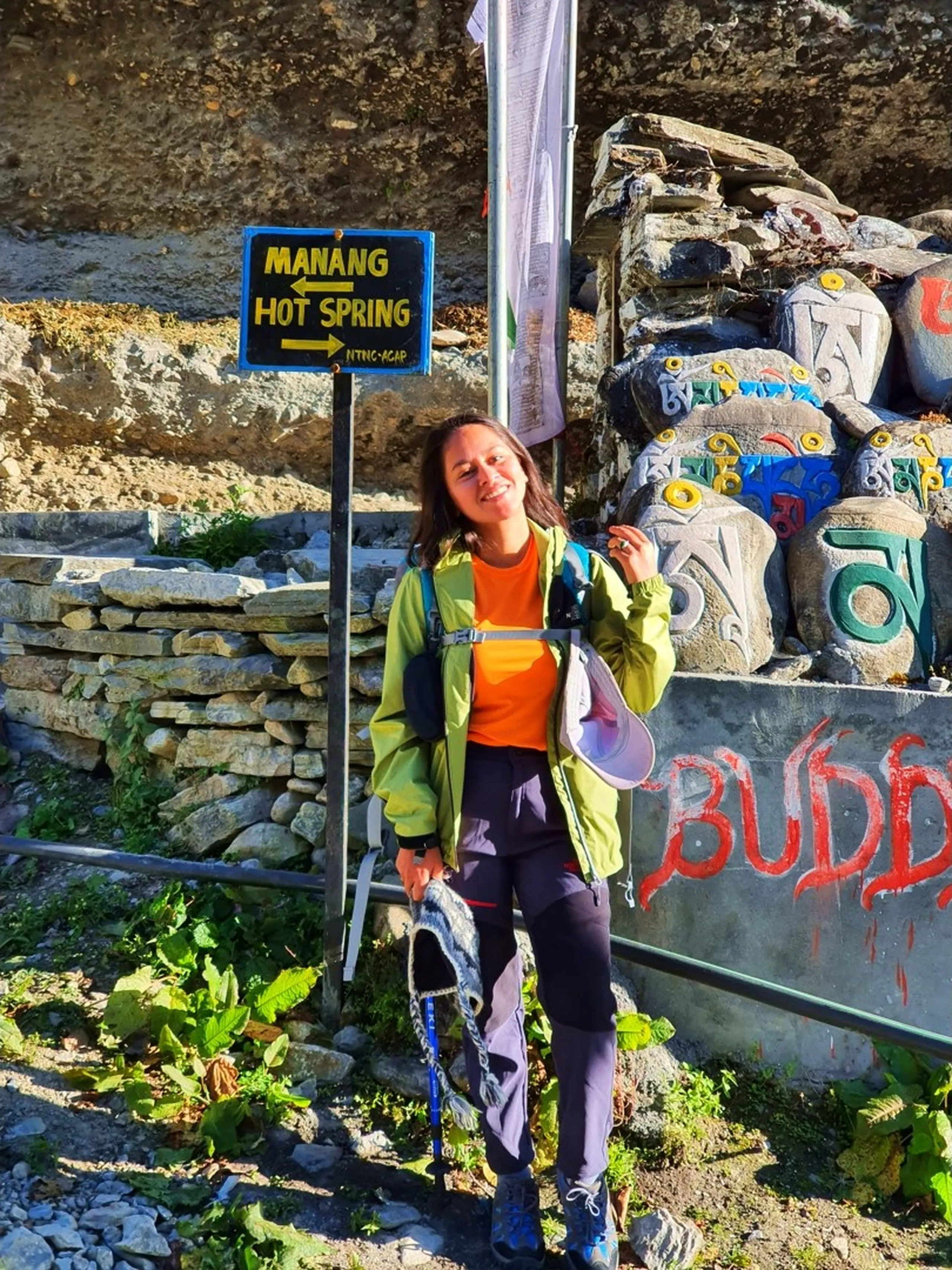 A young Nepali woman trekker smiles brightly while standing beside the 'Manang Hot Spring' directional signpost on the Annapurna Circuit trail - wearing a neon green trekking jacket over an orange top, dark trekking trousers, blue trail shoes, and carrying a trekking pole and a colorful Nepali wool hat - a pile of painted Mani prayer stones with Tibetan script in vivid colors stacked behind the sign - a white prayer flag overhead and 'BUDD' graffiti in red on a wall - rocky mountain cliffside in the background - an energetic and adventurous solo female trekker portrait at the Manang Hot Spring trail junction on the Annapurna Conservation Area trekking circuit, Nepal - great for Nepal solo female trekking blog content