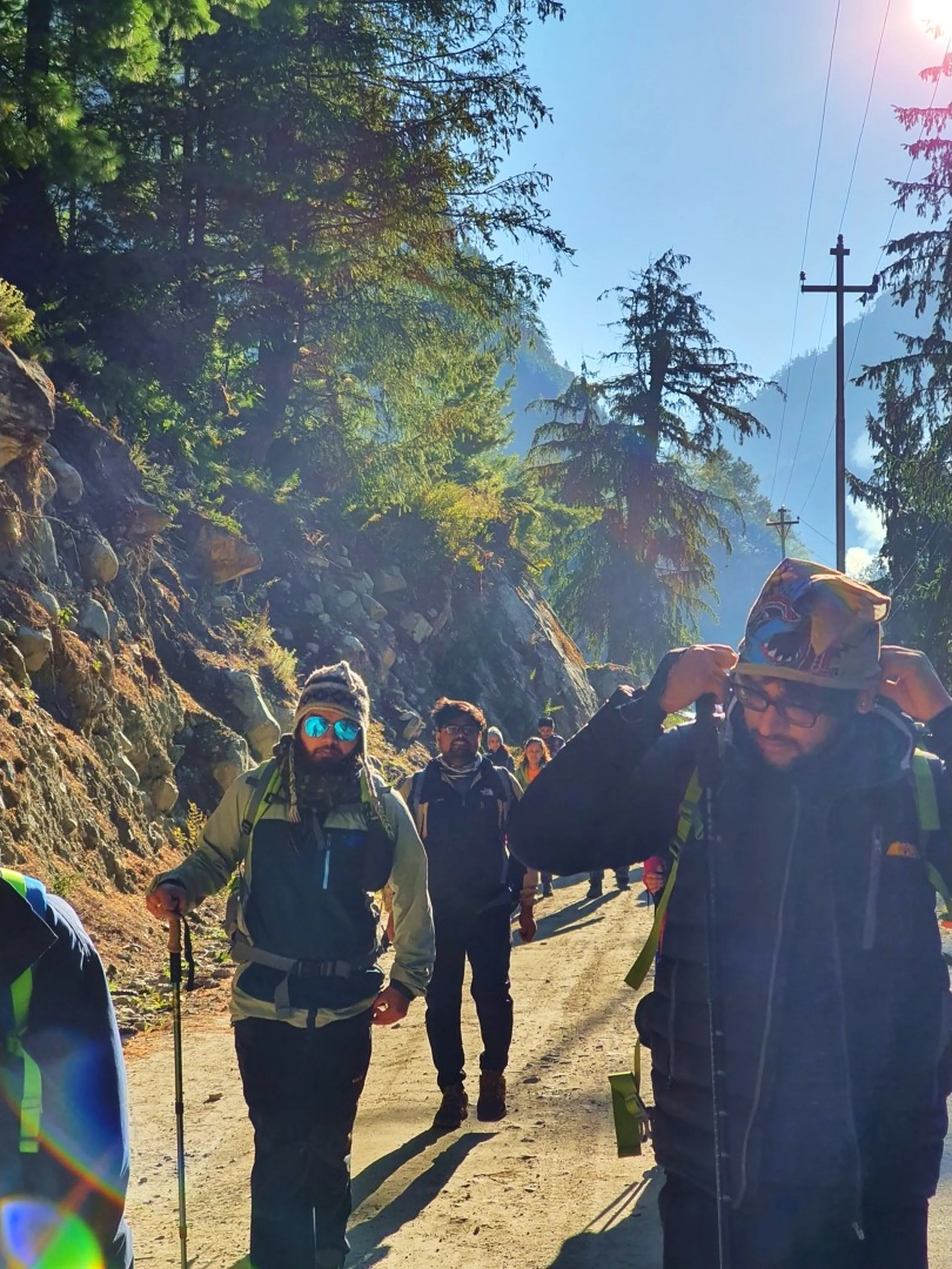 A candid action photograph of a group of Nepali trekkers walking along a sun-dappled pine forest mountain road on the Annapurna Circuit in Manang, Nepal - one in a green jacket with blue mirrored sunglasses leading with a trekking pole, another in all-black mid-frame, a third adjusting their wool hat at the edge - large boulders and scree slope on the left, tall pine trees filtering bright golden morning sunlight from the right - a long lens flare streak of rainbow light visible in the lower left - other trekkers visible further down the trail - a vivid and atmospheric Nepal Himalayan trekking trail photograph capturing the journey on the Annapurna Circuit to Manang, Ice Lake, and beyond
