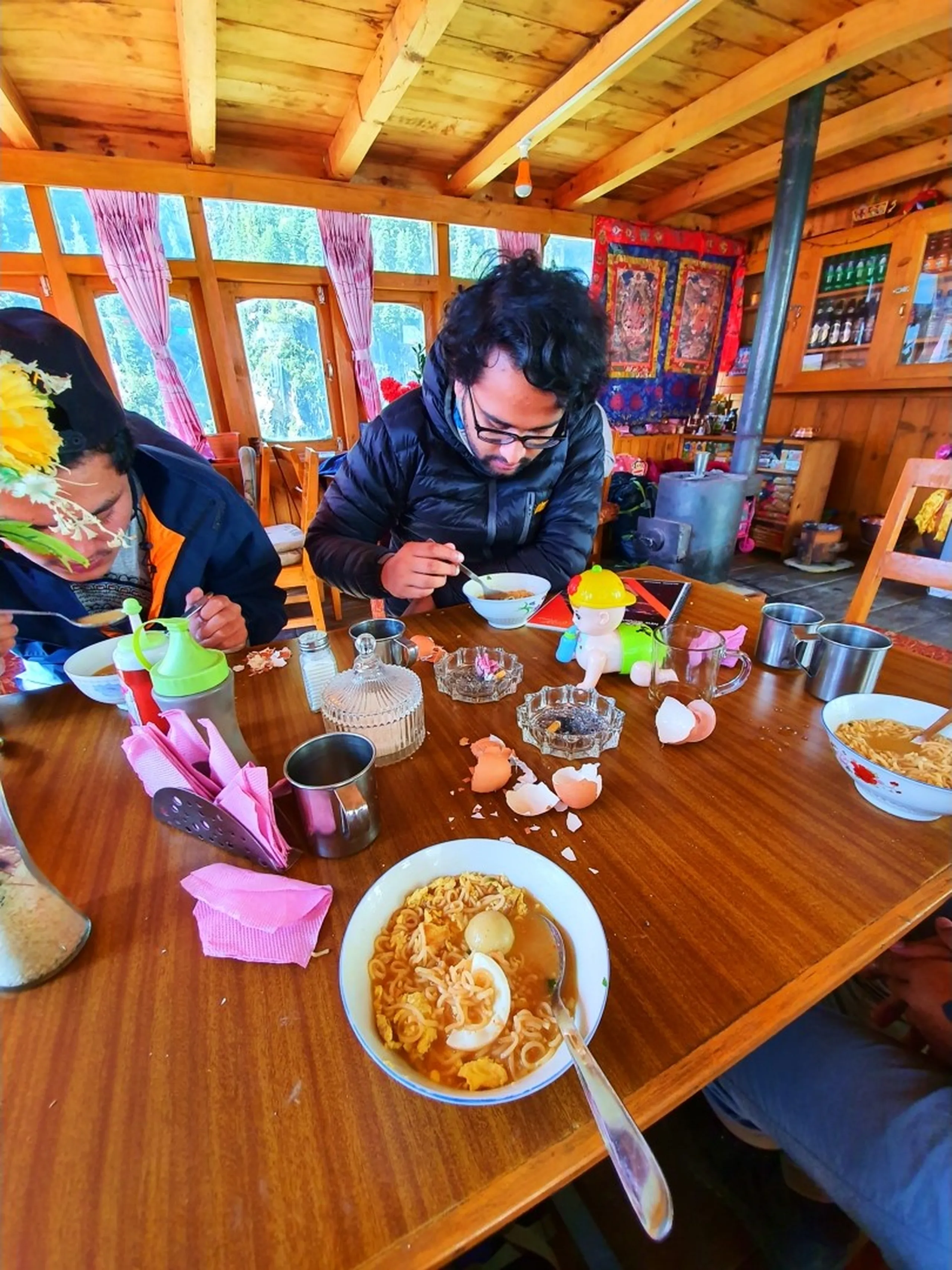A wide-angle interior shot of trekkers eating lunch inside a cozy wooden teahouse dining room on the Annapurna Circuit trail, Nepal - a young male trekker with glasses and black puffer jacket hunches over a bowl of steaming Wai Wai noodle soup with a boiled egg - another trekker in orange visible to the left eating - the rustic pine-paneled room is warm and inviting with wooden beam ceilings, pink curtains, a metal wood-burning stove, and a traditional Tibetan thangka painting on the back wall - a glass cabinet stocked with drinks and supplies - the table scattered with empty eggshell halves, condiments, steel mugs, and a plastic cartoon figurine - an authentic Nepal mountain teahouse meal photograph, capturing the essential noodle soup lunch culture of Annapurna Circuit trekking