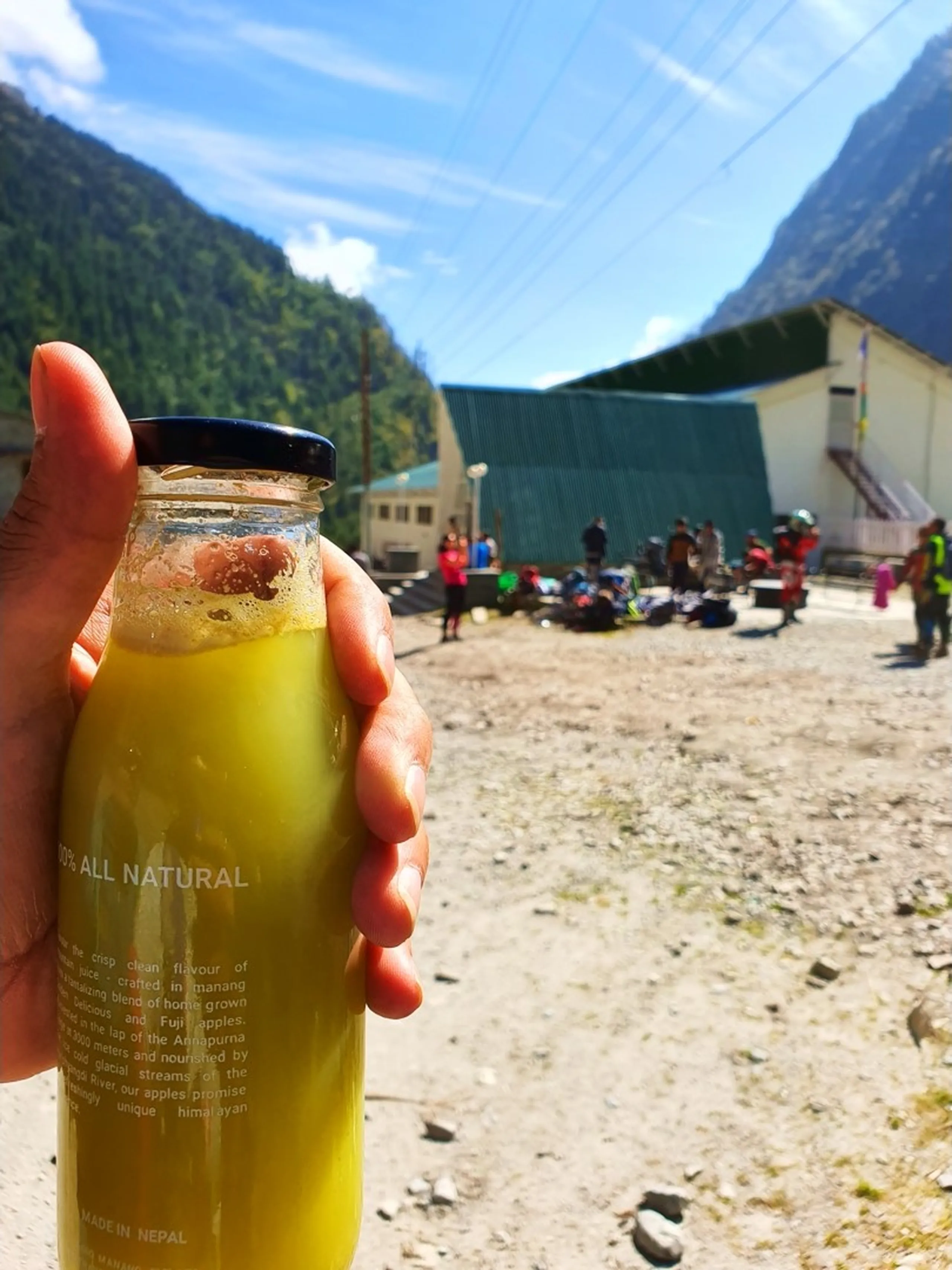 A hand holds up a glass bottle of bright golden-yellow fresh apple juice labeled '100% All Natural - Made in Nepal - crafted in Manang from home-grown Fuji apples nourished by the Annapurna glacial streams' - in the blurred background, a group of trekkers rest on the rocky ground outside a green-roofed building in Bhratang village, Manang, Nepal - dramatic Himalayan valley walls visible on both sides under a blue sky - a refreshing and distinctive Nepal Himalayan local produce photograph, showcasing the famous Manang apple juice from Agro Manang Pvt Ltd farmhouse in Bhratang - perfect for Nepal trekking food and local produce travel blog content