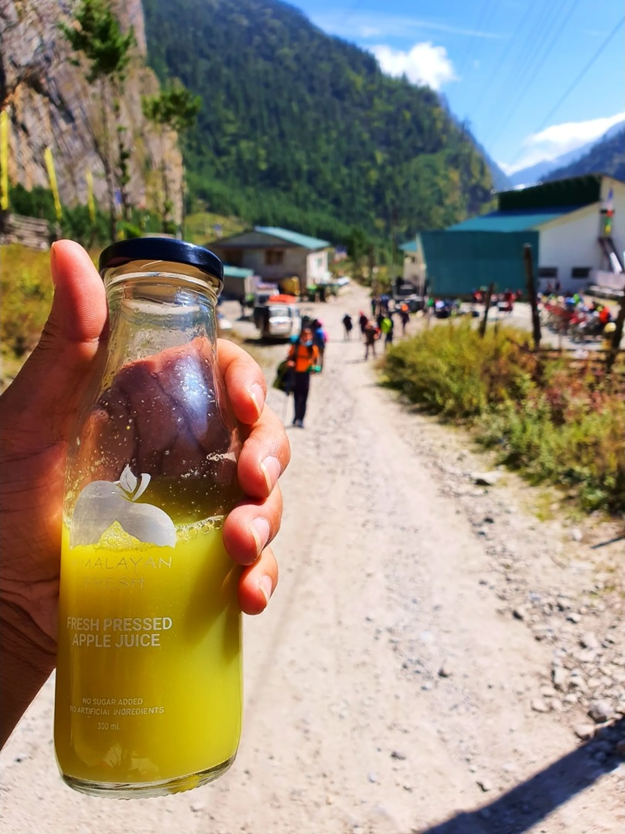 A close-up of a hand holding a glass bottle of vibrant yellow 'Himalayan Fresh Pressed Apple Juice - No Sugar Added, No Artificial Ingredients, 300ml - Made in Nepal' - labeled with a white apple motif - in the soft-focus background, a group of loaded trekkers walk along the dirt trail road past green-roofed buildings in Bhratang, Manang, Nepal - dramatic Himalayan gorge cliffs visible in the background under a bright blue sky - cyclists and other trekkers visible in the distance - a compelling Nepal local food and trekking trail photograph, featuring the iconic fresh-pressed apple juice produced at the Agro Manang farmhouse in Bhratang on the Annapurna Circuit, a must-try for all trekkers passing through Manang