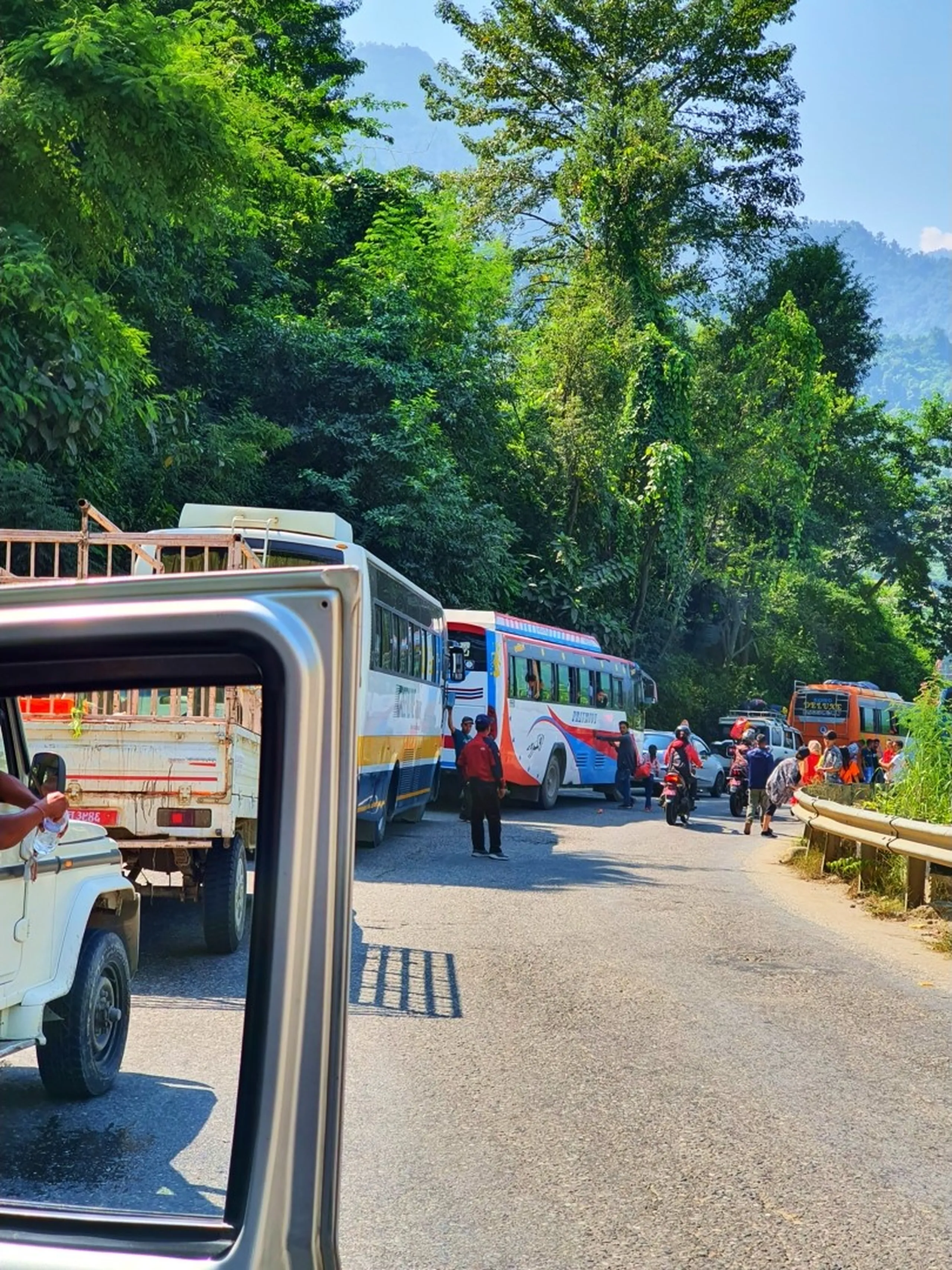 A long traffic jam on a narrow mountain highway in Nepal photographed from inside a vehicle window, showing a convoy of colorful tourist buses, trucks, and jeeps at a standstill surrounded by dense subtropical jungle and green hills - bystanders visible on the roadside - a typical Nepal highway traffic experience on the route to Manang and the Annapurna Circuit, showing the reality of Nepal mountain road travel