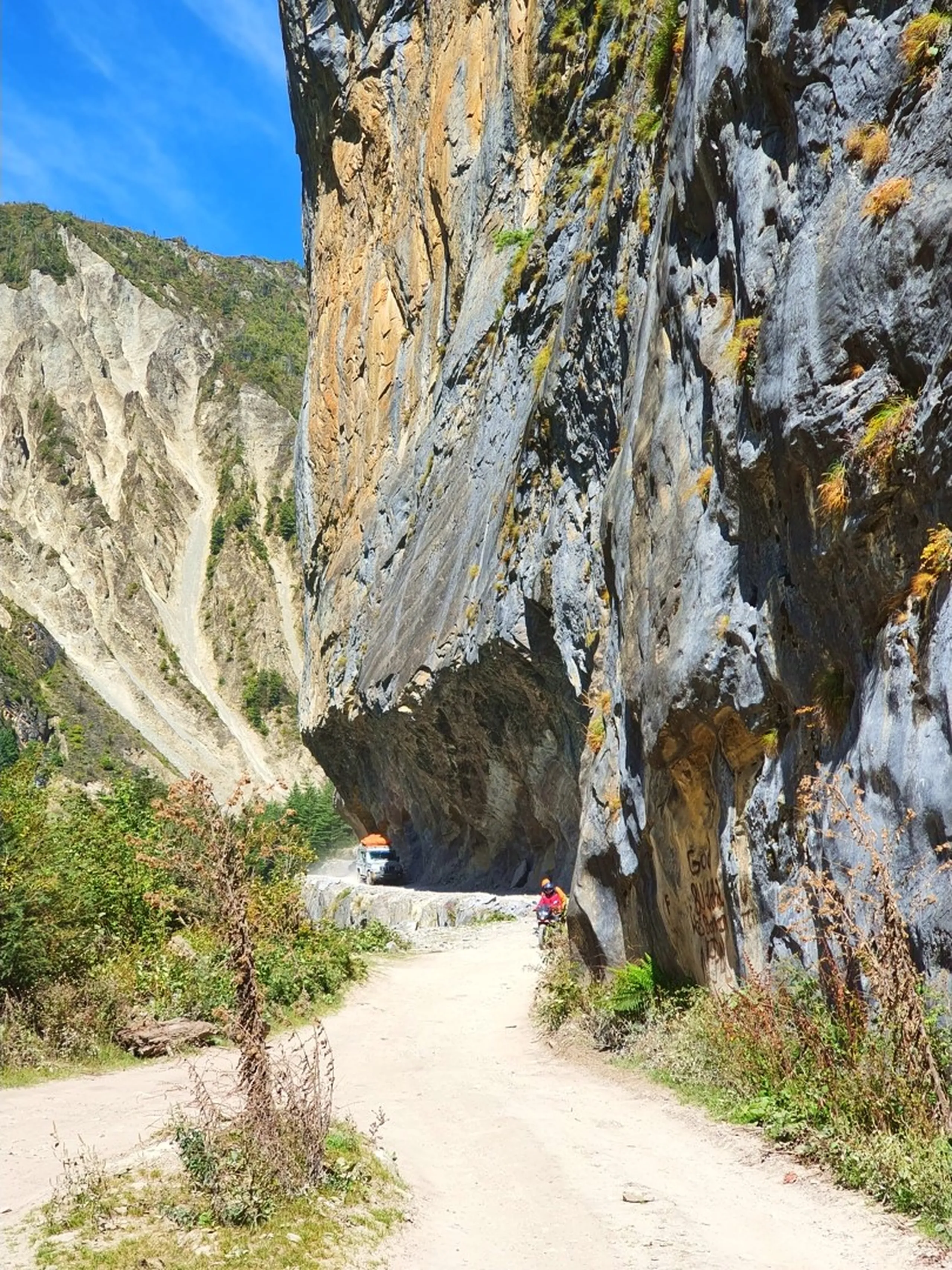 A dramatic wide-angle landscape photograph of a narrow trekking trail road carved along the base of a towering overhanging cliff face in Manang, Nepal - the sheer rock wall rises vertically from the road, its surface streaked in grey, yellow, and rust tones with patches of shrub clinging to crevices - a small bicycle rider in an orange jacket and another figure barely visible beneath the overhang give scale to the enormous cliff - pine forest and eroded scree slopes visible across the valley - brilliant blue sky visible in the upper left corner - a breathtaking and dramatic Nepal mountain trail photograph on the Annapurna Circuit between Chame and Pisang, showcasing the extraordinary geological formations and raw terrain of trekking to Manang