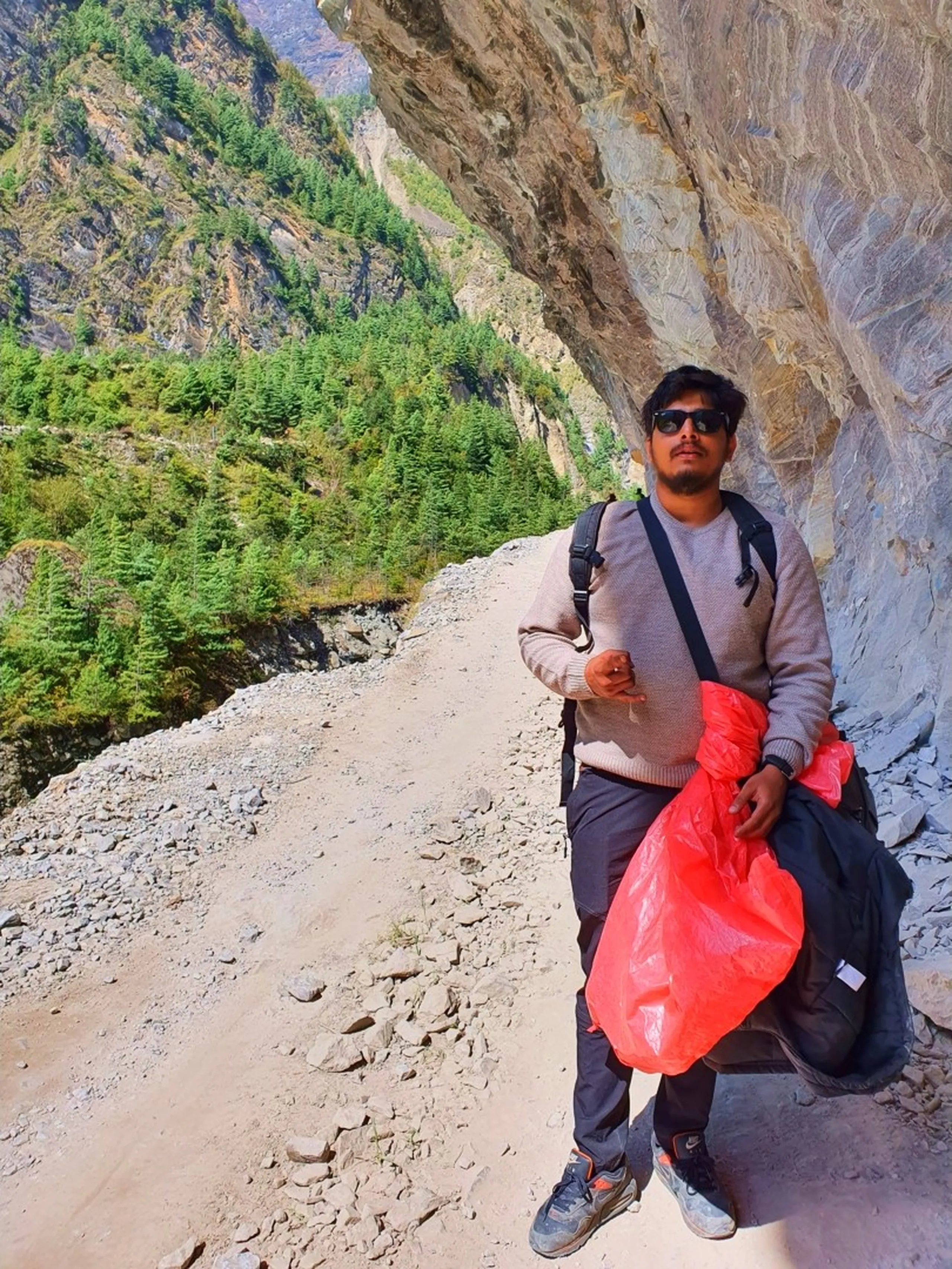 A young male Nepali trekker walks along a narrow dirt trekking trail carved beneath a dramatic overhanging rock cliff in Manang, Nepal - wearing a dusty rose-beige sweater, dark trekking trousers, Nike Air Max sneakers, and dark sunglasses - carrying a crossbody bag and two bright red plastic bags - the trail narrows between the cliff wall on the right and a steep rocky drop to a pine-forested valley on the left - a candid and adventurous Nepal trekking trail portrait under a natural rock overhang on the Annapurna Circuit route between Chame and Pisang, showcasing the dramatic terrain of the trail to Manang, Nepal