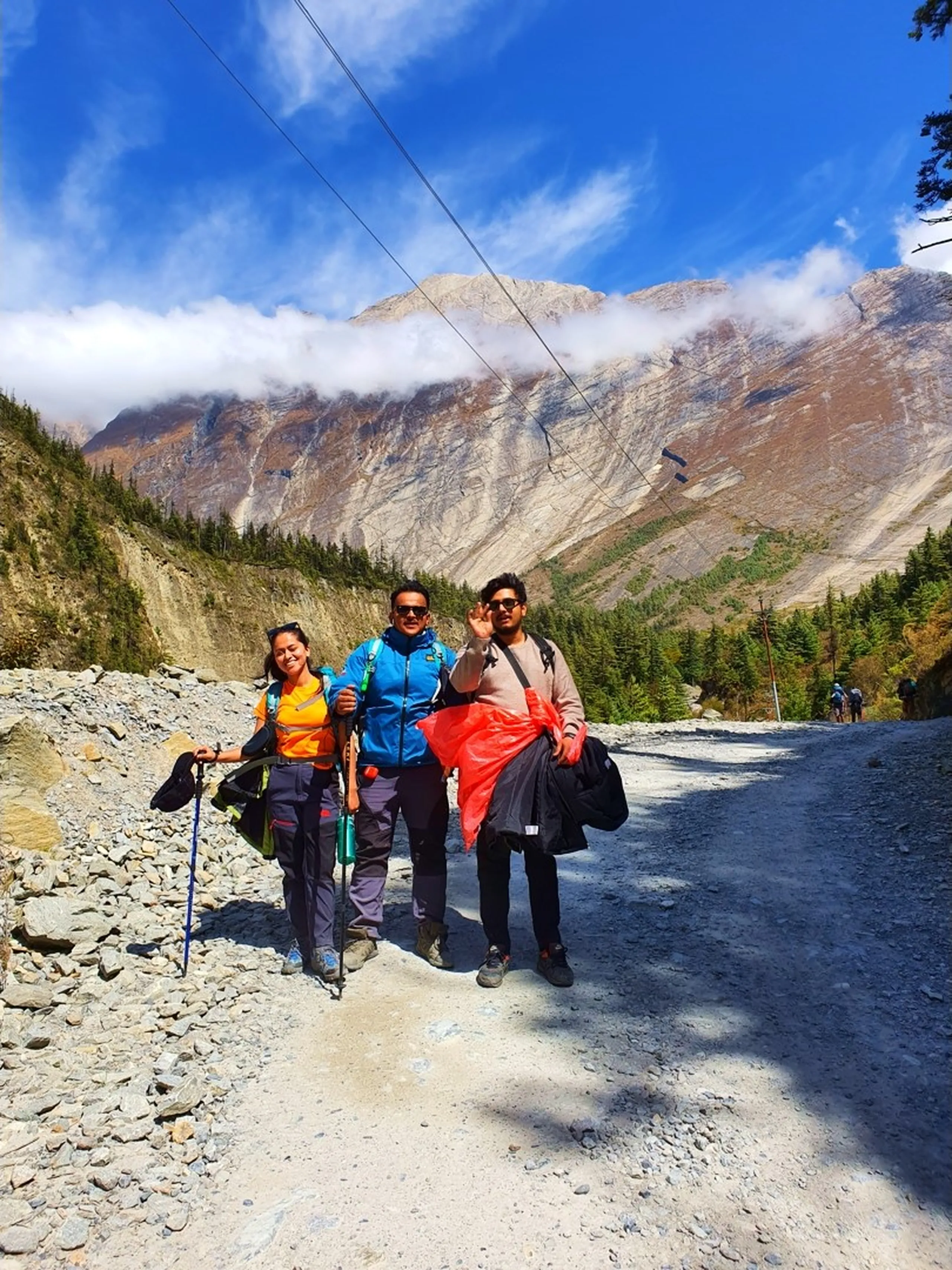 Three young Nepali trekkers pause and pose together on a wide rocky mountain trail in Manang district, Nepal - a young woman in an orange top, green jacket, trekking trousers, and blue shoes holds a trekking pole on the left - a man in a blue trekking jacket and grey trousers with a trekking pole stands in the center giving an 'OK' hand gesture - another in a beige sweater with red plastic bags stands on the right - a massive grey and rust-striped Himalayan cliff face with white cloud bands and patches of pine forest fills the entire background under a vivid blue sky - other trekkers visible on the trail behind them - a vibrant Nepal Annapurna Circuit trekking trail group portrait with dramatic high-altitude mountain scenery near Pisang, Manang